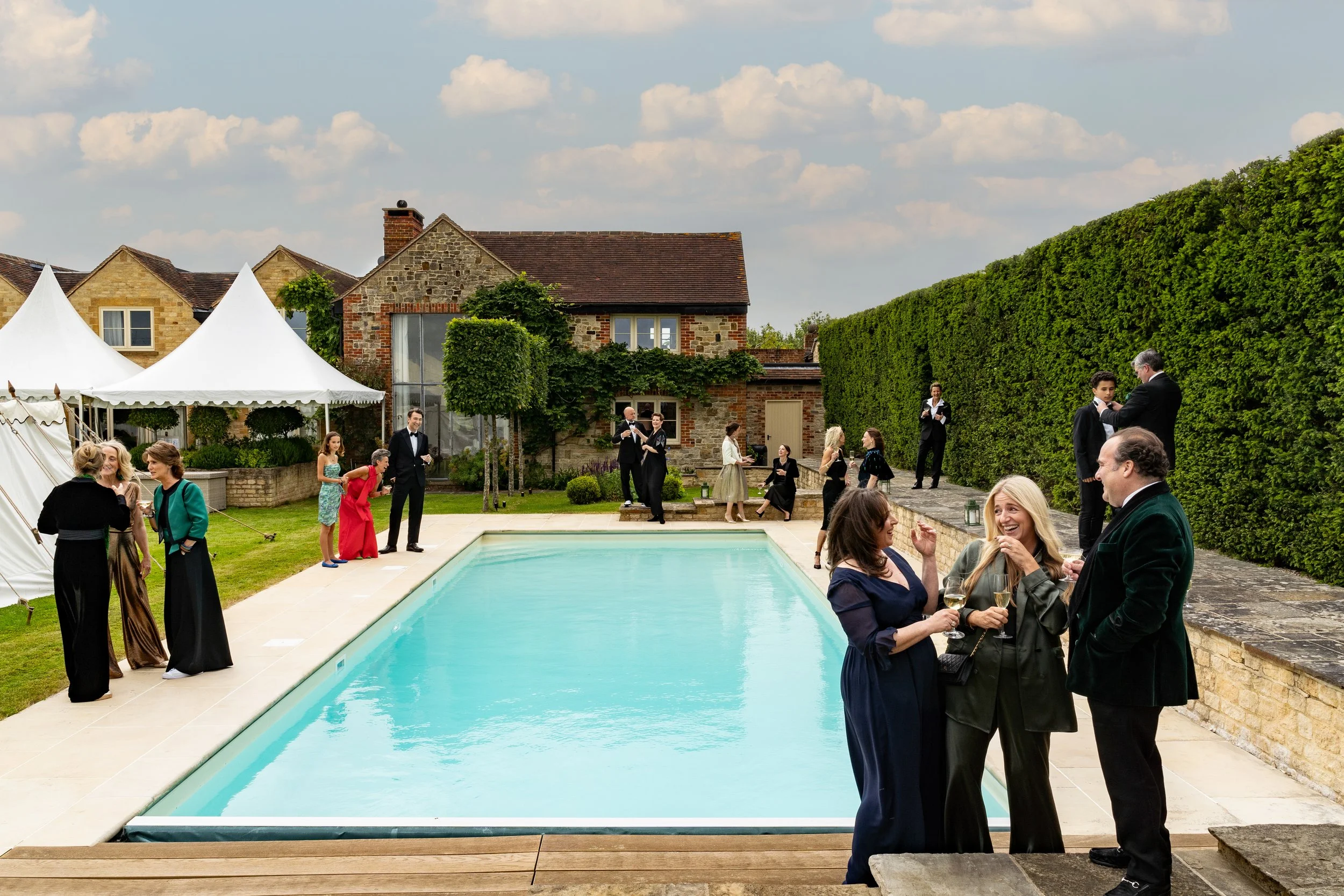 Guests socializing by a swimming pool at an outdoor event, with traditional stone houses, tents, and a tall hedge in the background.