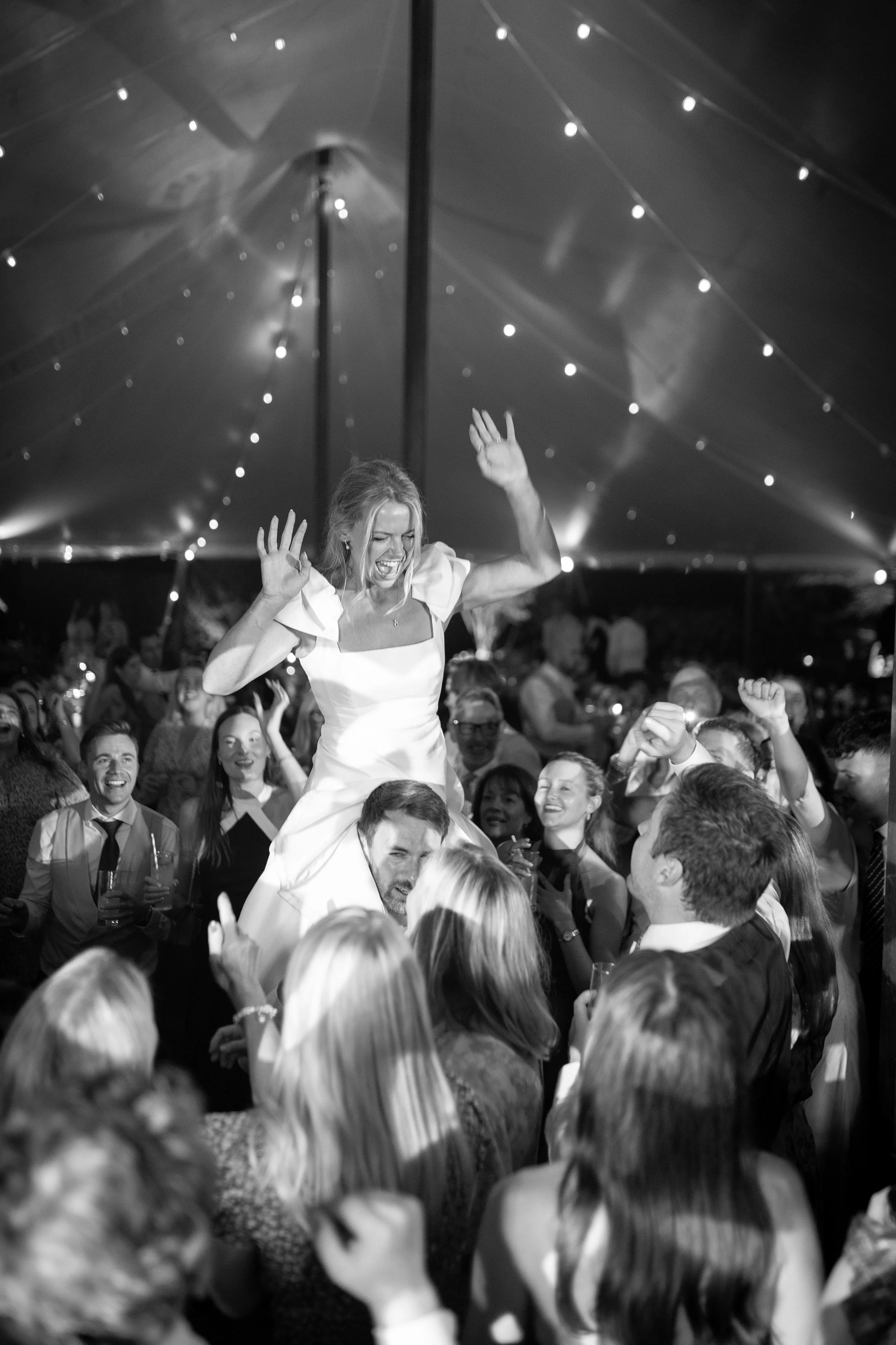 A woman in a white dress is being lifted by a man in a crowd under a decorated tent at a celebration, with people smiling and raising their hands.