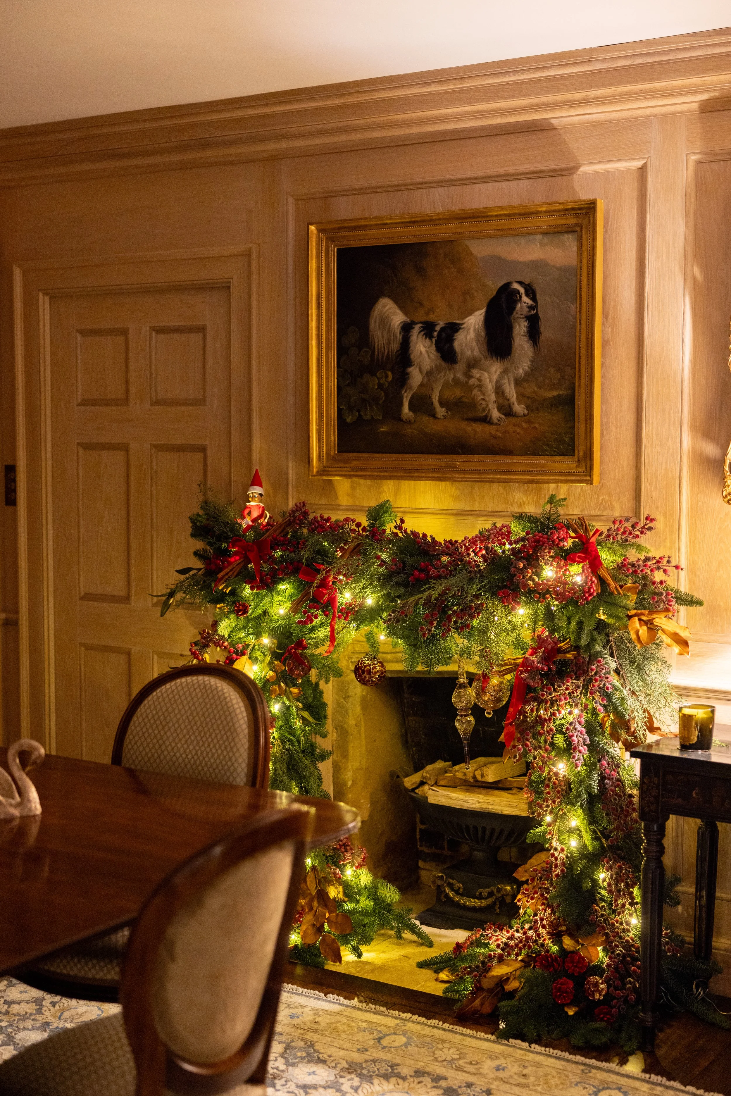 Festively decorated fireplace with greenery, red berries, ribbons, and ornaments, topped with a small Santa figurine, in a warm, wood-paneled room with a framed dog portrait overhead.