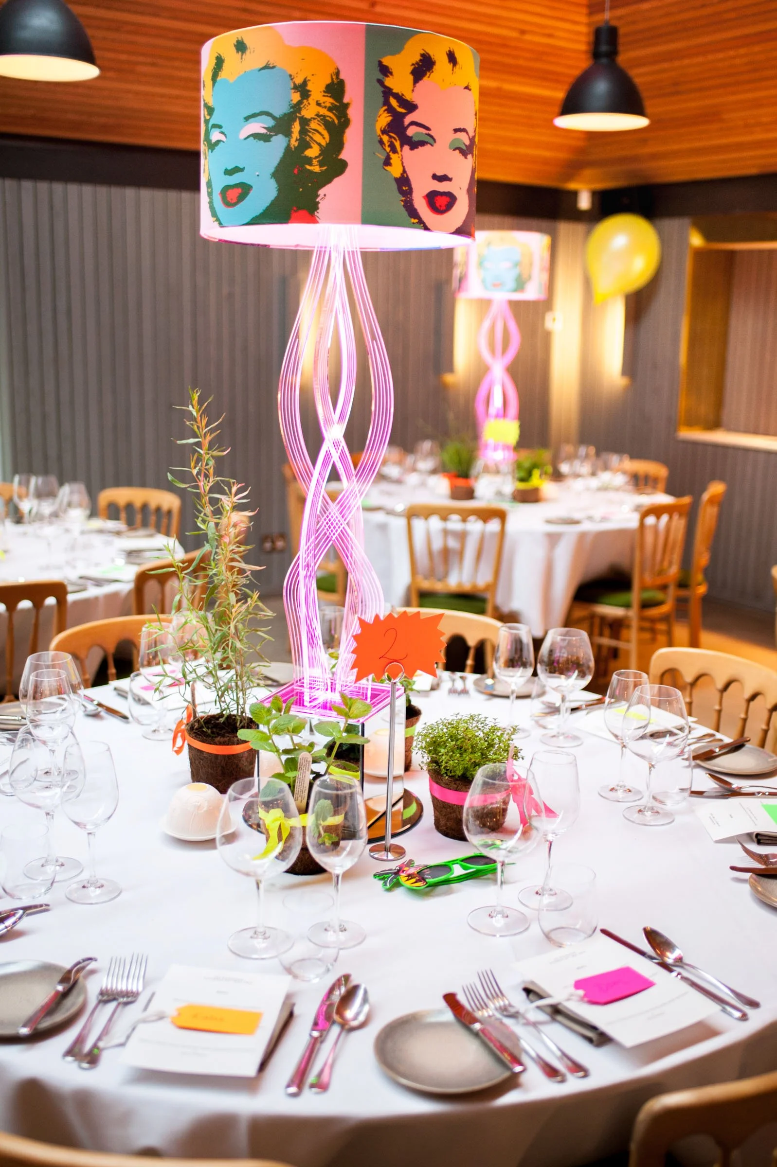 A decorated round banquet table set for an event, featuring multiple wine glasses, silverware, and place cards across a white tablecloth. A central pink neon-style lamp with an image of Marilyn Monroe's face in a pop art style, with a lit candle and 