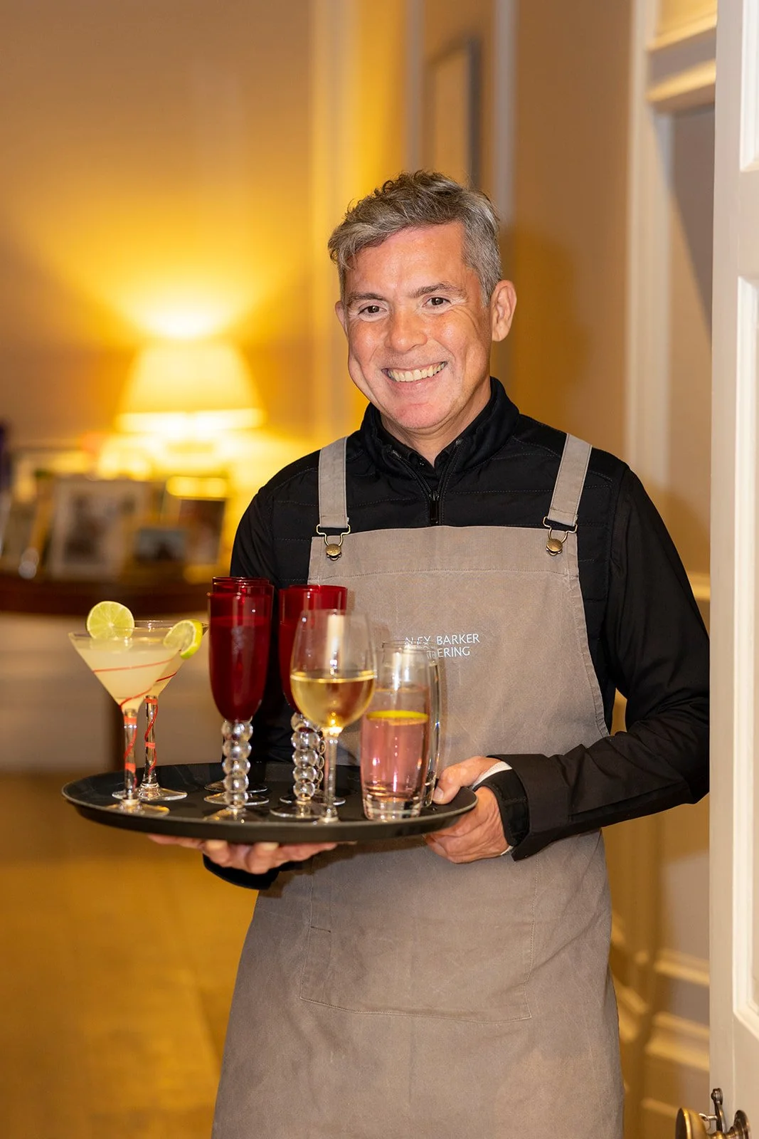 A smiling man in a black shirt and beige apron holding a black tray with various cocktails in a warmly lit room