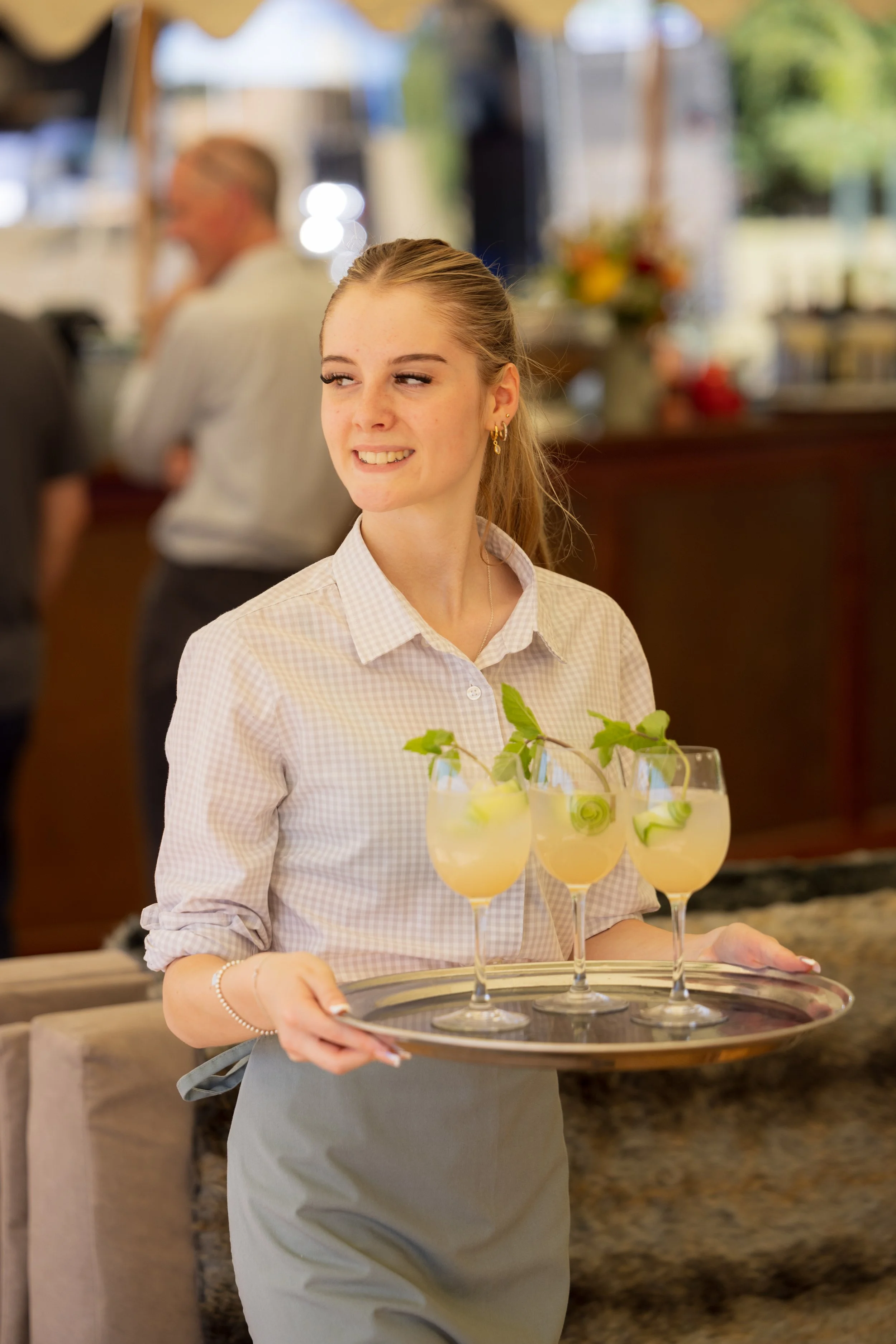 A young female server holding a tray with three glasses of lemonade garnished with lemon slices and mint leaves, inside a restaurant or cafe.