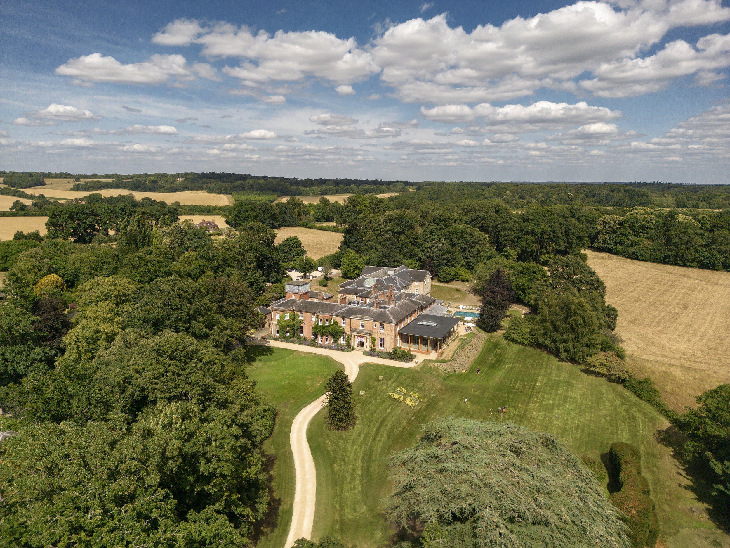 A large estate with a mansion surrounded by trees and lush greenery, with fields and a cloudy sky in the background.