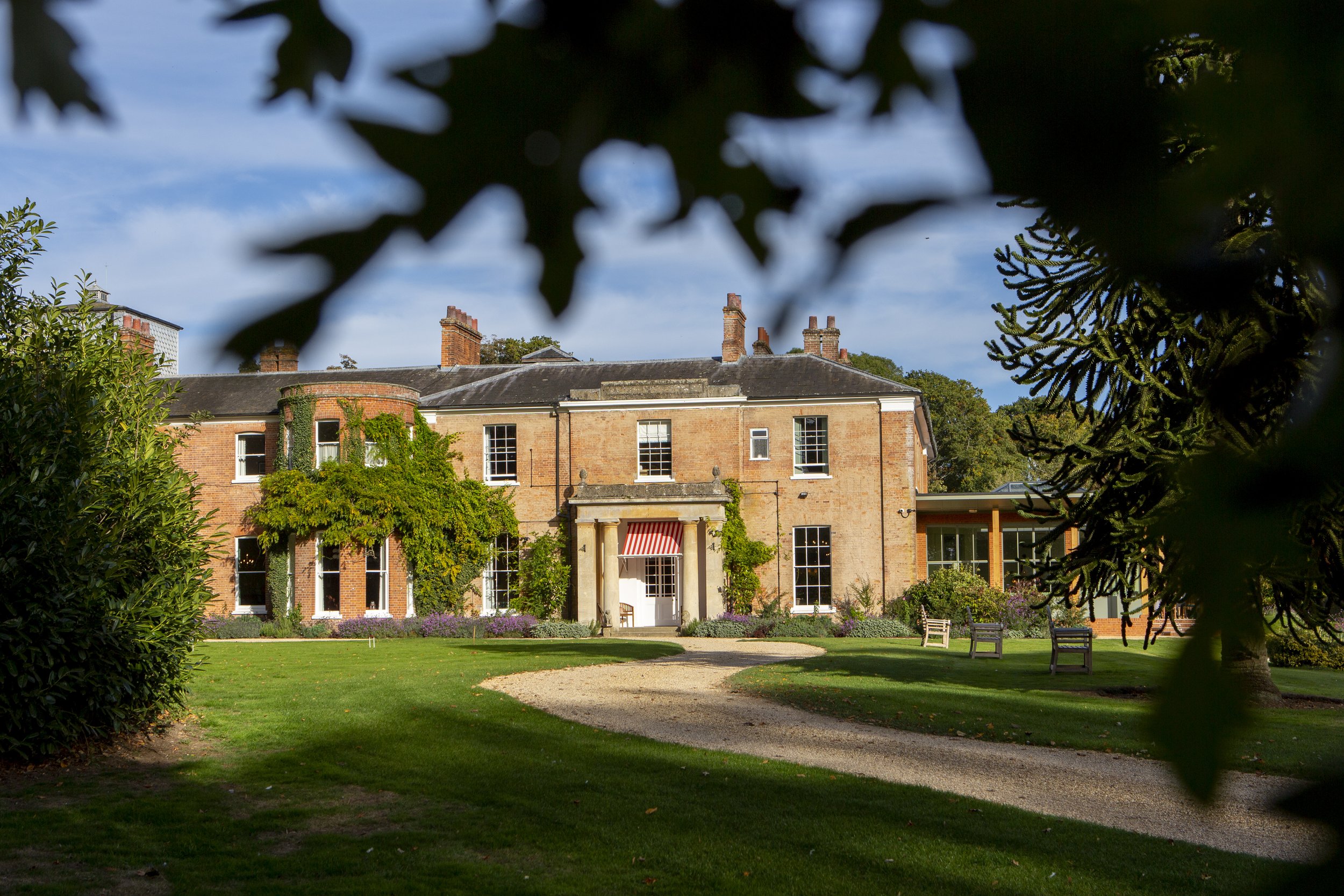 A large historic brick house with multiple chimneys, surrounded by well-manicured lawn, benches, and trees, viewed through a frame of leaves in the foreground.