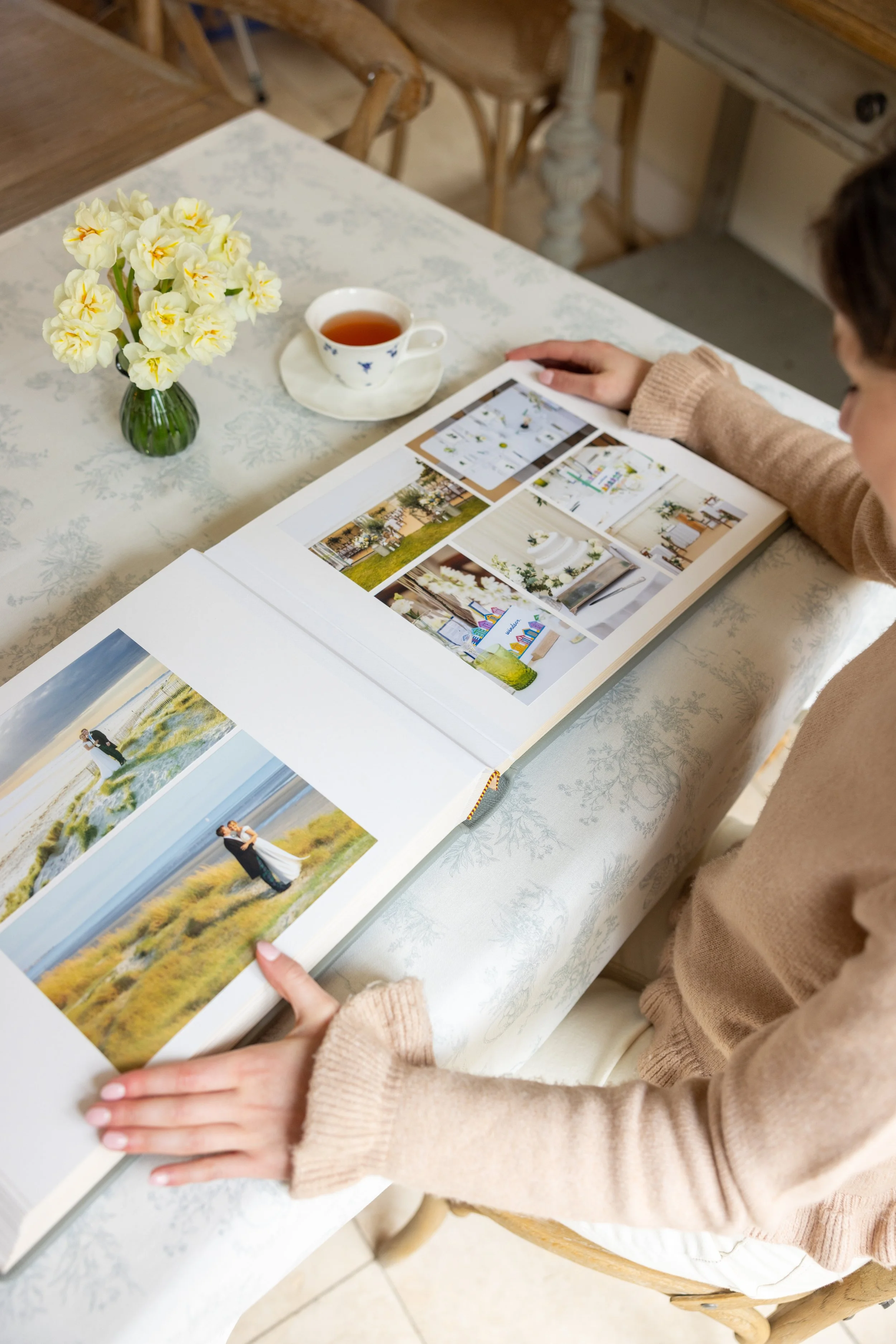 Person looking at a photo album with pictures of wedding scenes on a table with a cup of tea and a vase of yellow flowers nearby.
