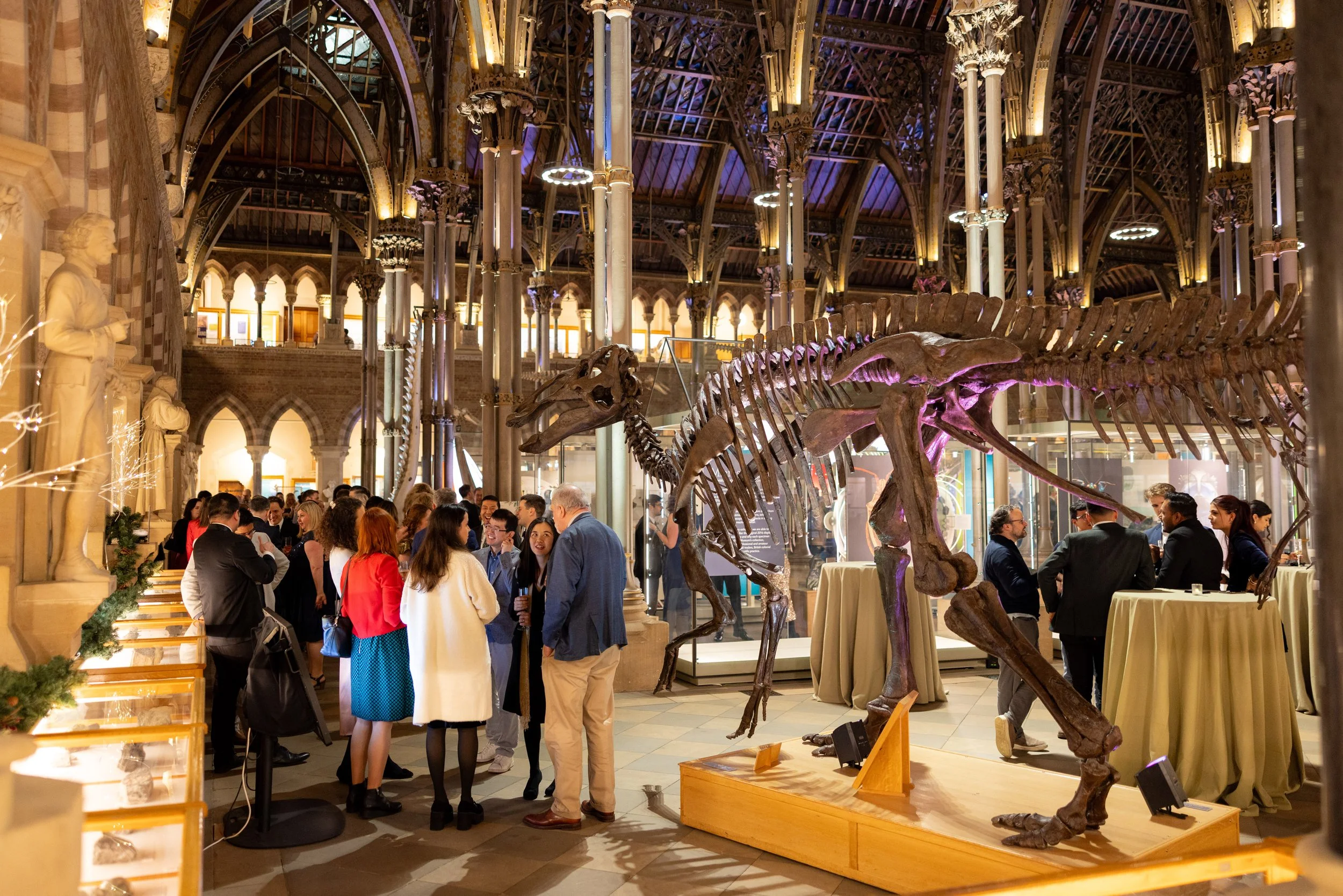 A group of people gathered in a museum's dinosaur exhibit, featuring a large dinosaur skeleton, ornate architectural details, and statues along the walls.