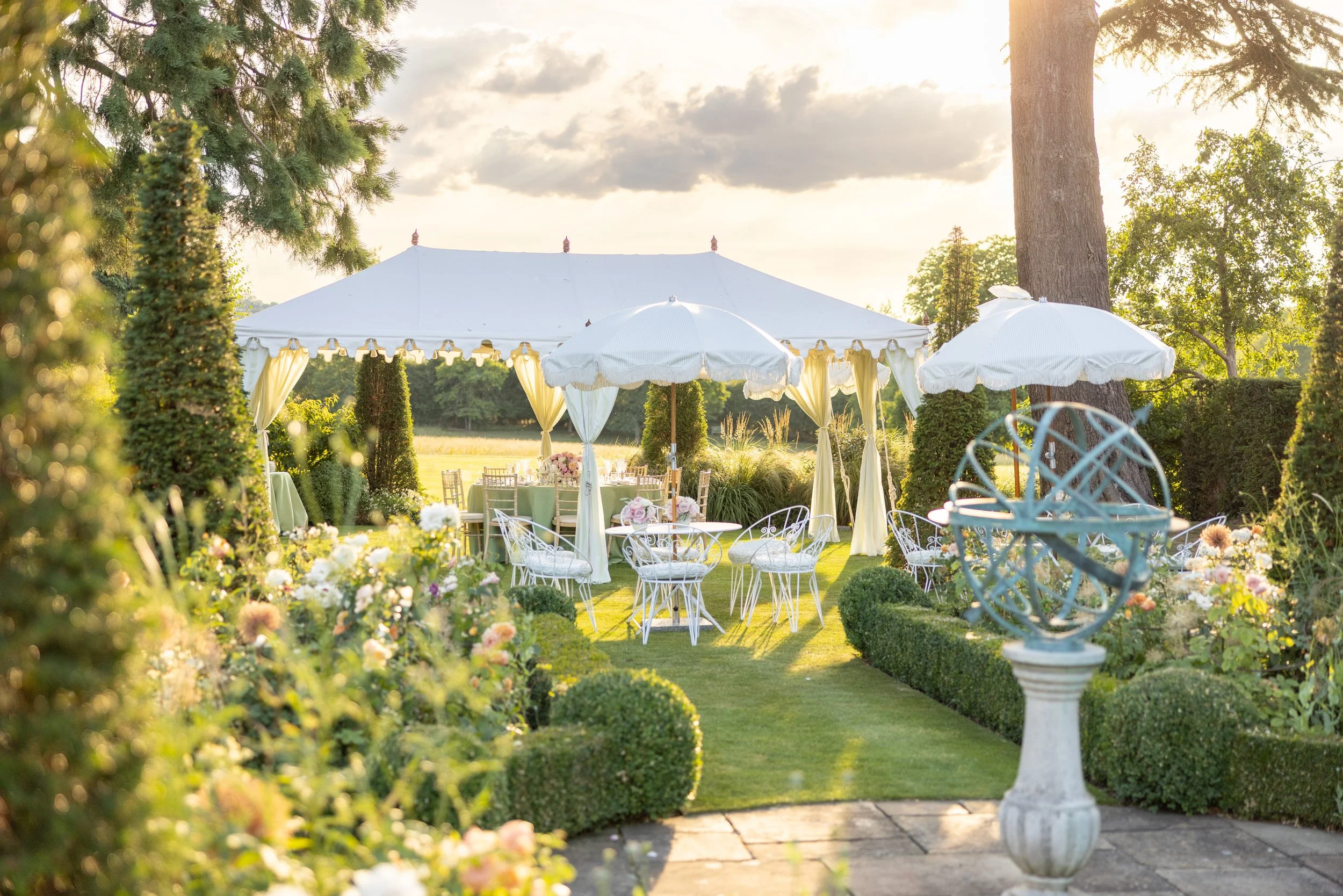 Elegant outdoor garden setup with white tables, chairs, umbrellas, and a large white canopy tent, surrounded by lush green plants and flowers, during sunset.
