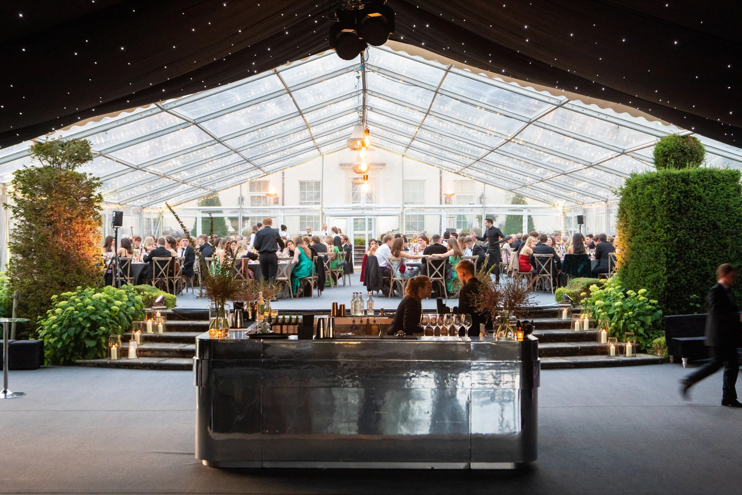 A large group of people seated at tables inside a glass tent for a formal event, with waitstaff attending to guests, and a bar area in the foreground. The tent is decorated with string lights and lush greenery.