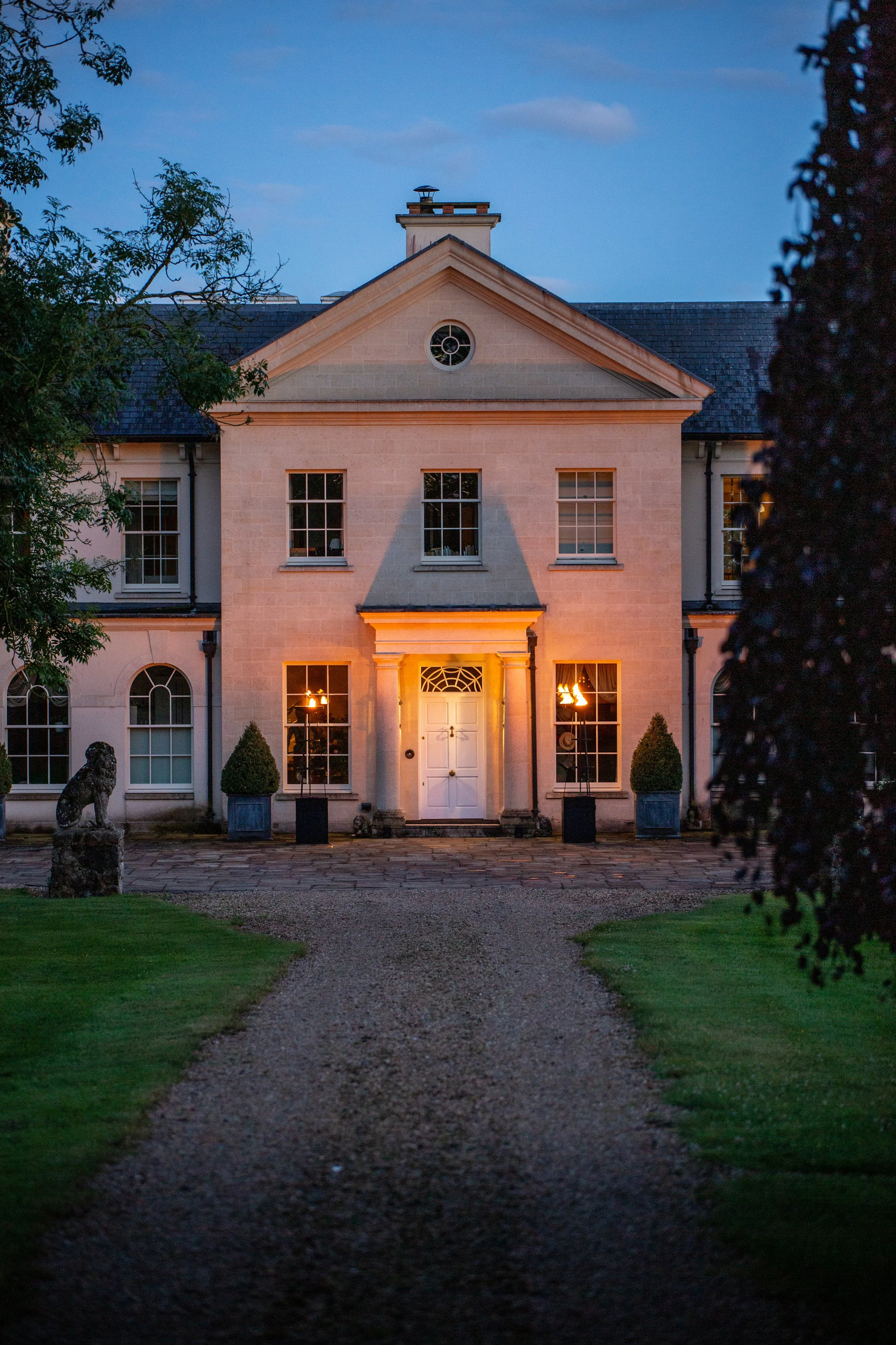 Front view of a large, elegant house illuminated at dusk, with a gravel driveway leading to the white front door, flanked by two potted plants and lit lamps, surrounded by greenery and trees.