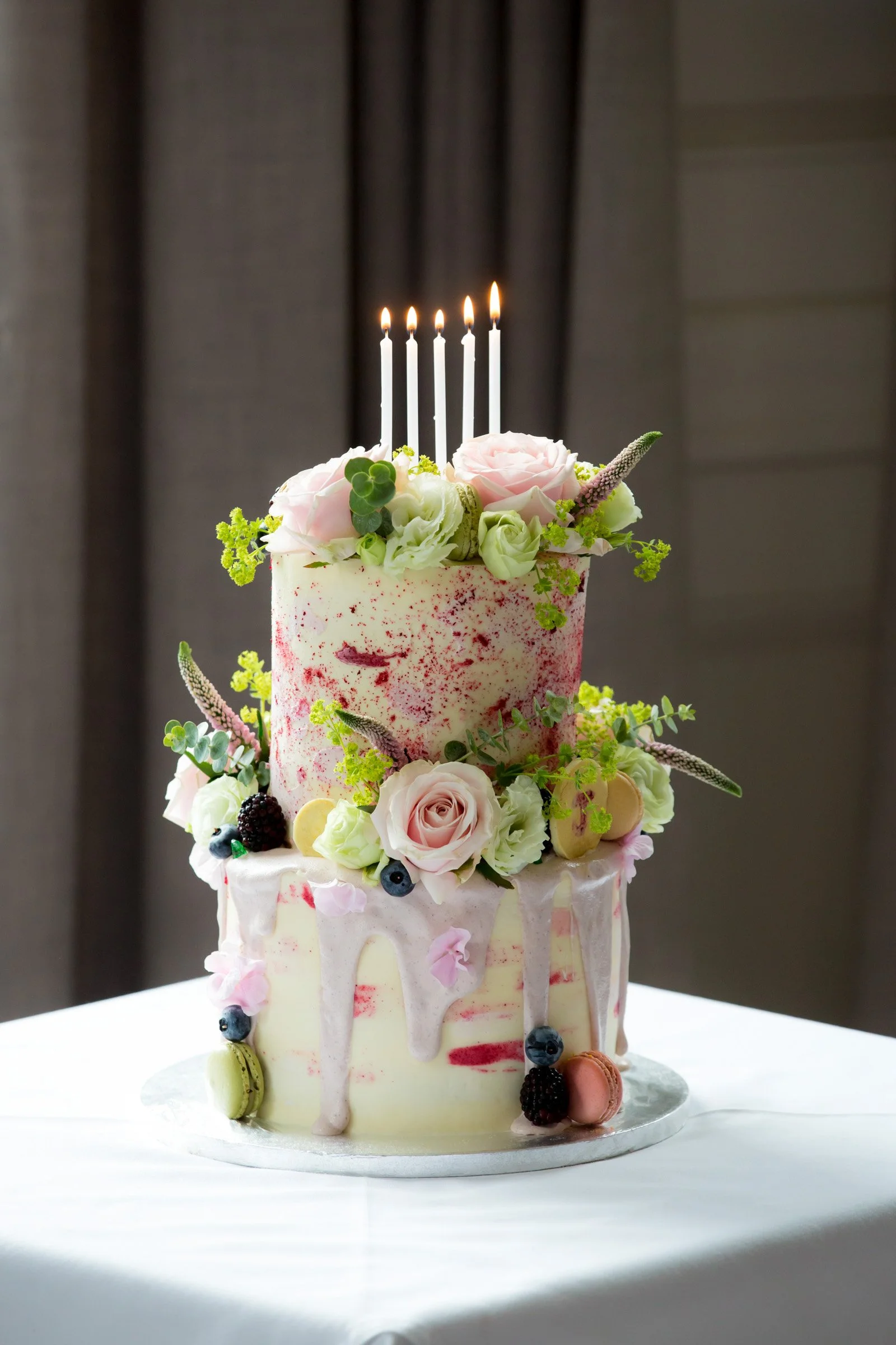Two-tiered wedding cake decorated with pink and white roses, blueberries, blackberries, macarons, and greenery, with five lit candles on top, on a white table.