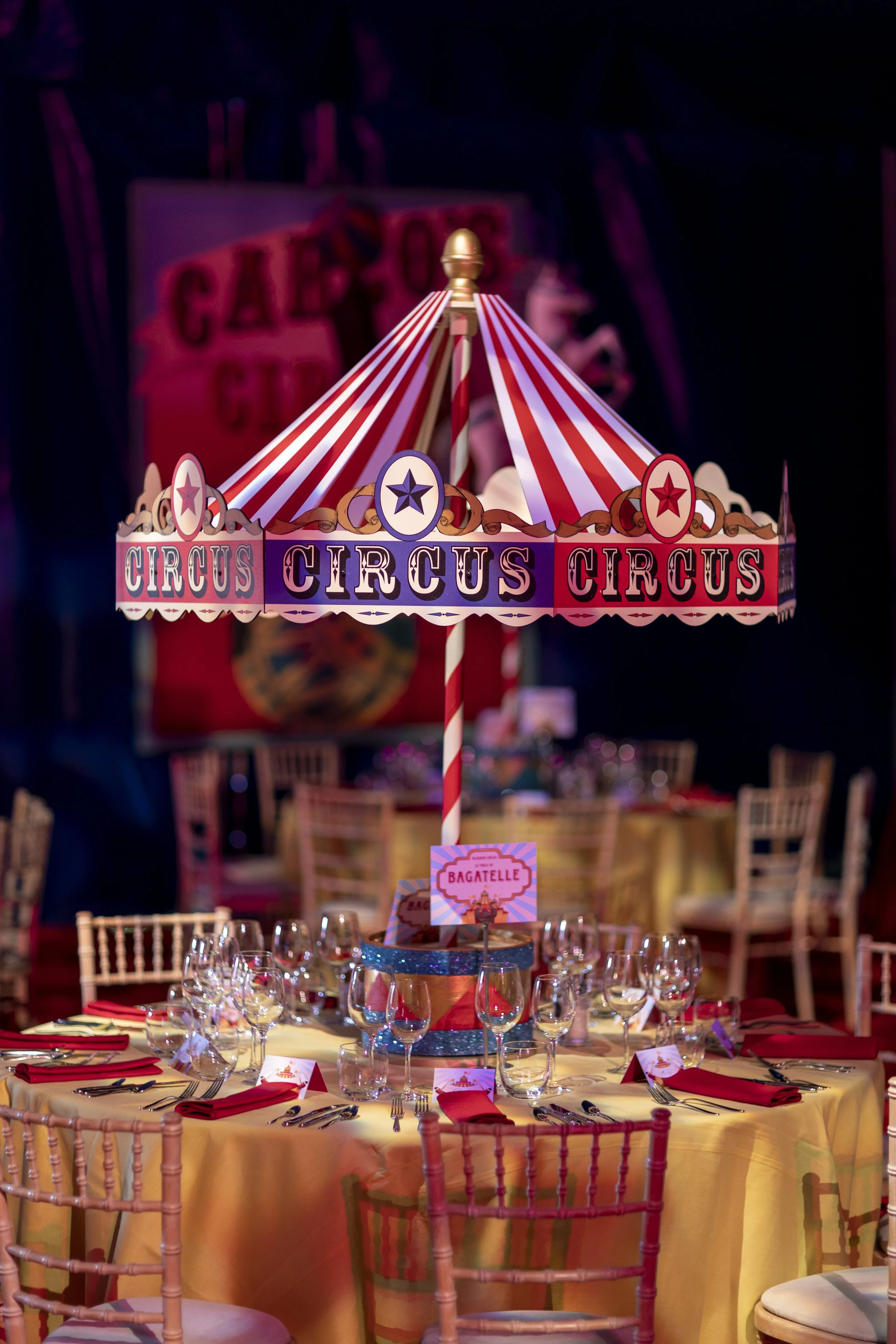 Circus-themed event table setting with a round yellow tablecloth, red napkins, wine glasses, silverware, and a centerpiece featuring a miniature circus tent with red, white, and blue stripes and the word "CIRCUS" repeated on the border.