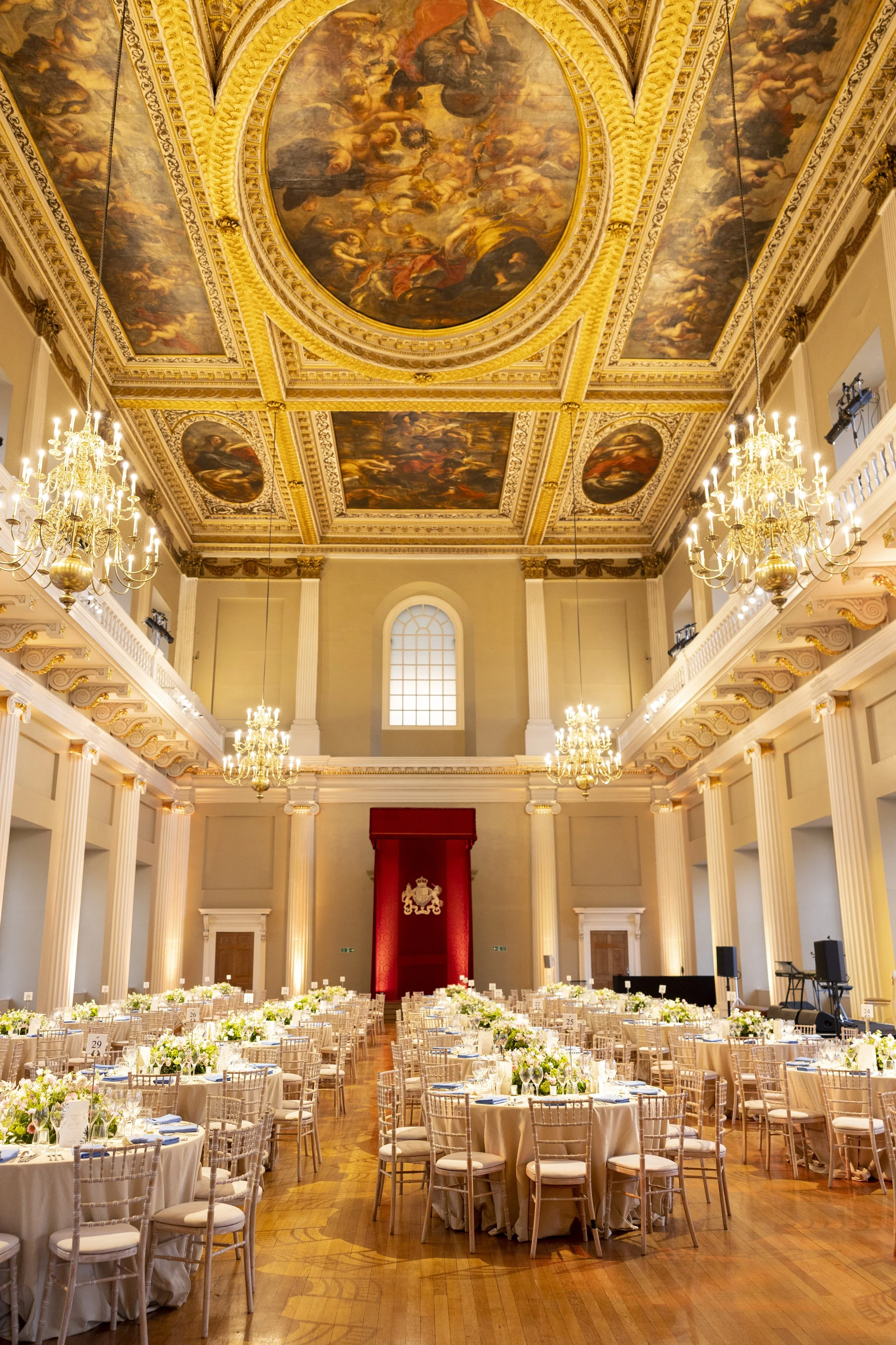 Elegant banquet hall with ornate ceiling paintings, chandeliers, and round guest tables decorated with floral centerpieces.