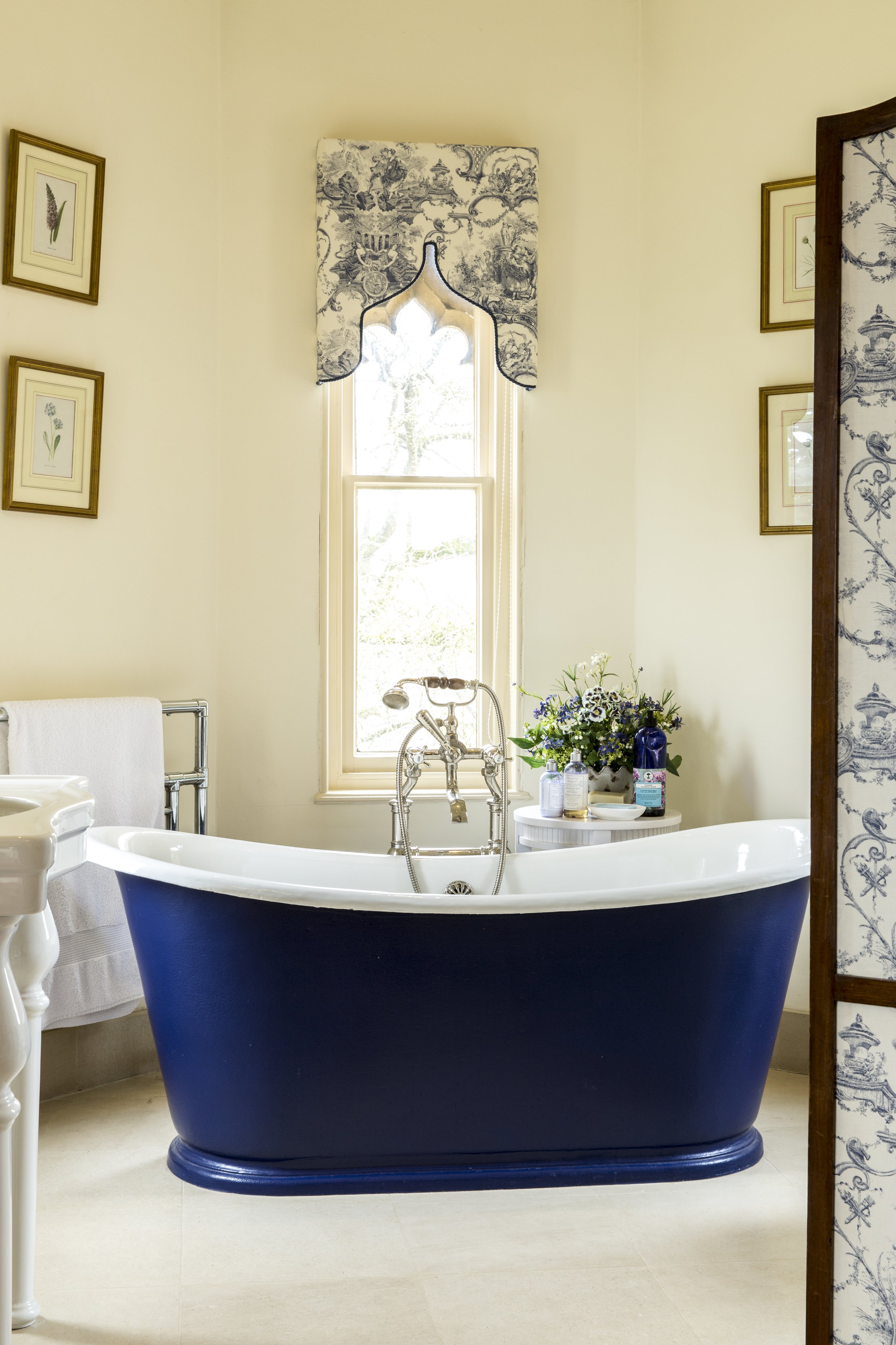 A vintage-style bathroom with a classic blue and white clawfoot bathtub near a window with blue and white patterned curtain. There are framed botanical prints on the wall and a small white side table with flowers and toiletries.