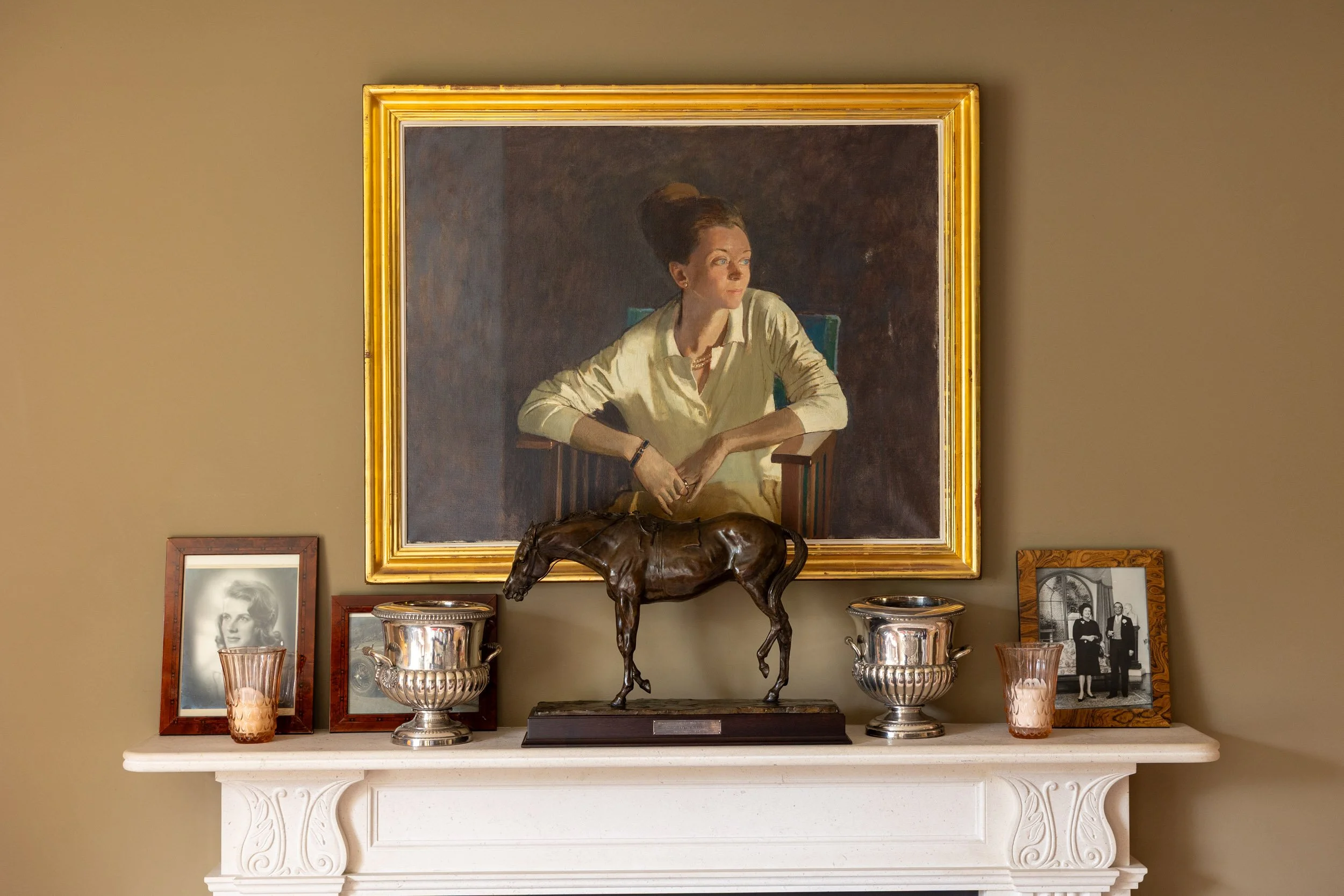 A mantelpiece decorated with framed photographs, a bronze horse sculpture, and silver vases in front of a large portrait painting of a woman in a cream blouse sitting with her arm resting on a chair.