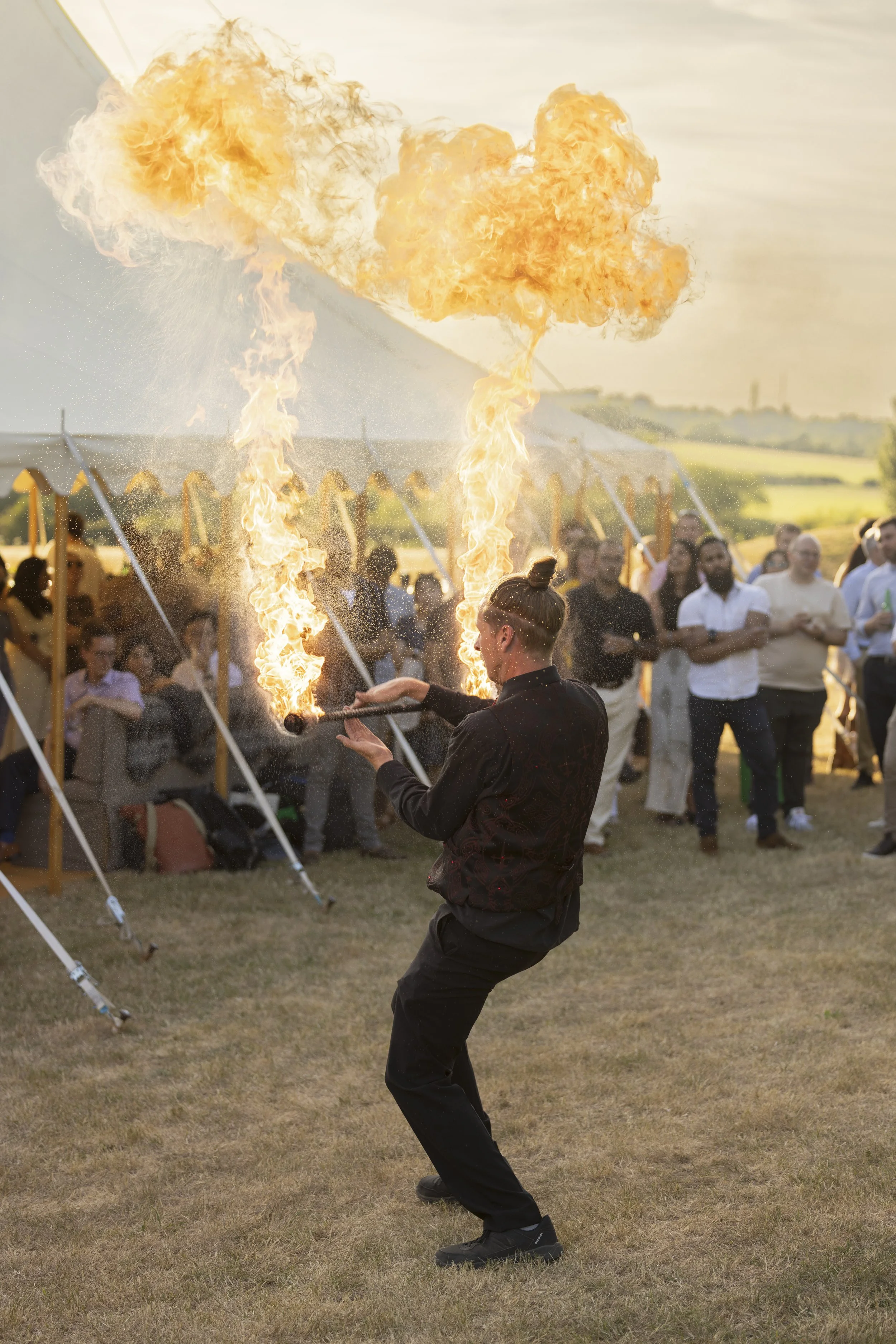 A performer doing a fire-breathing act outdoors in front of a crowd during the daytime, with flames shooting from their mouth into the air.