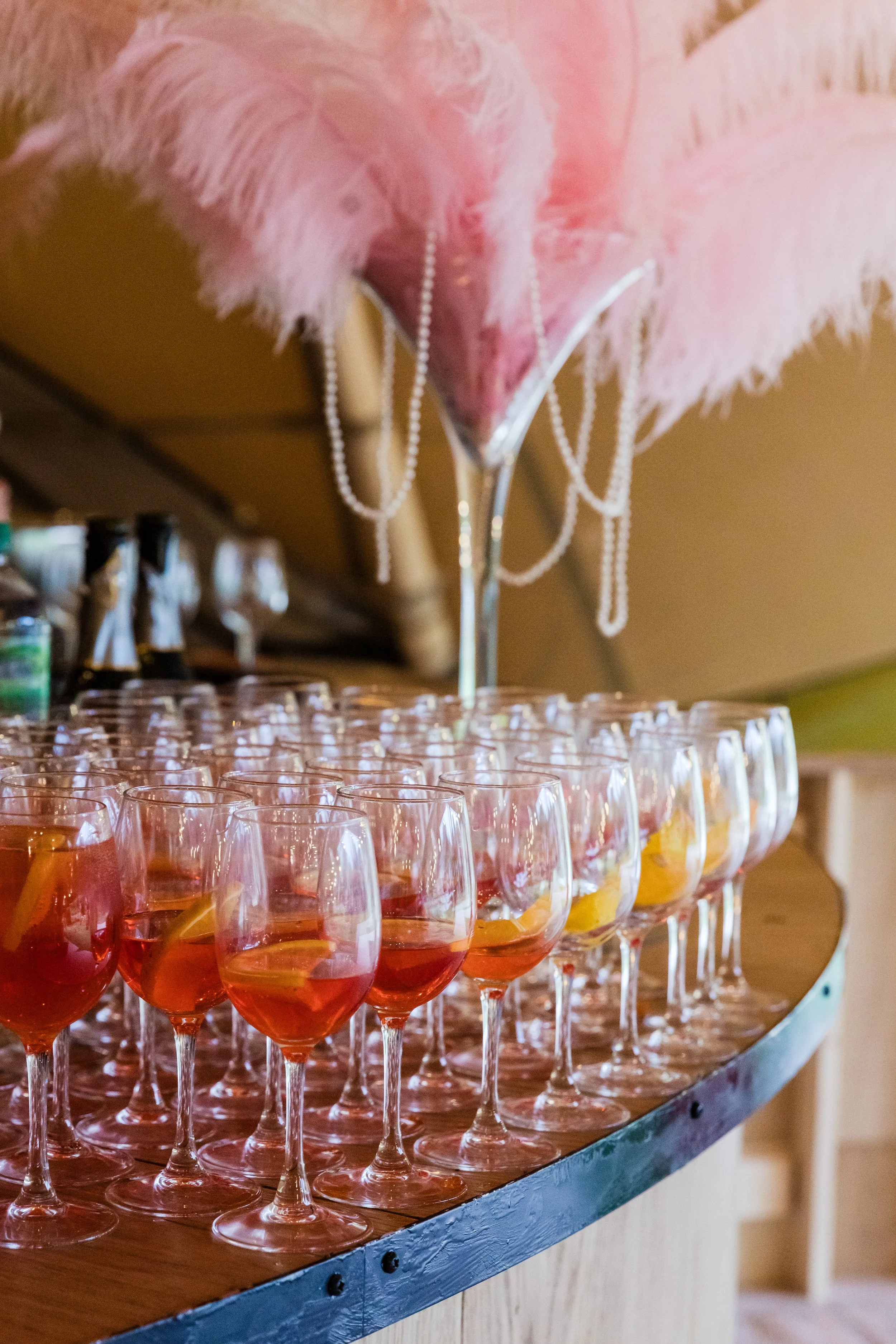 A large table with many glasses filled with a pink drink garnished with lemon slices. Pink feathers and string beads decoratively hang above the glasses.