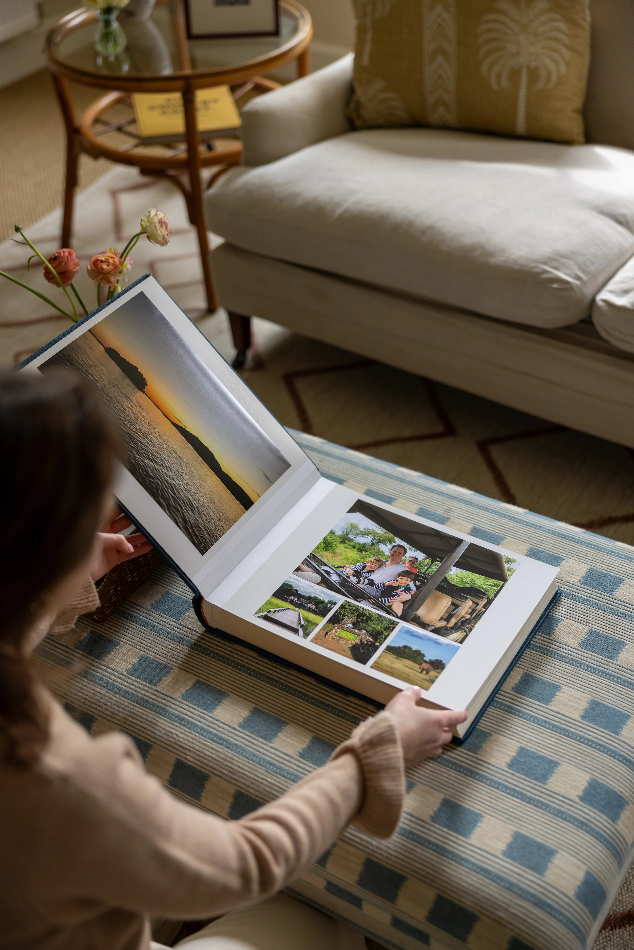 Person sitting on a striped blue and beige rug looking at a photo album with landscape and family pictures, next to a sofa with a blanket, a pillow, and a side table with a glass vase and books in a cozy living room.