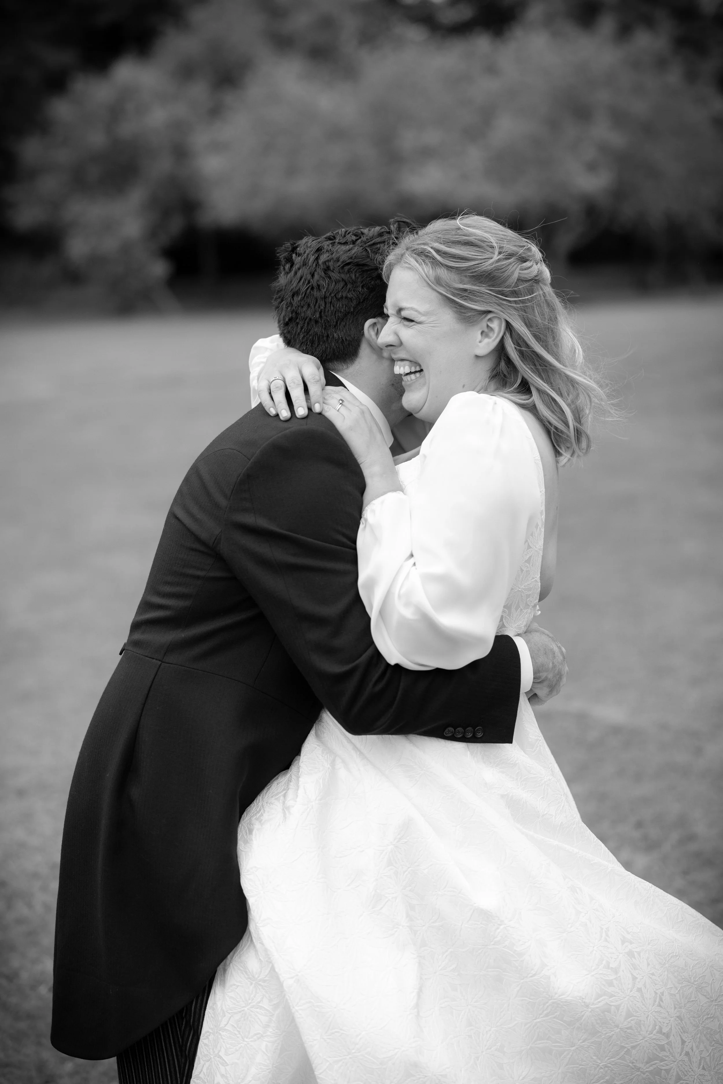 A black-and-white photo of a newlywed couple embracing outdoors, smiling and laughing, with the bride's eyes closed and the groom's face hidden.