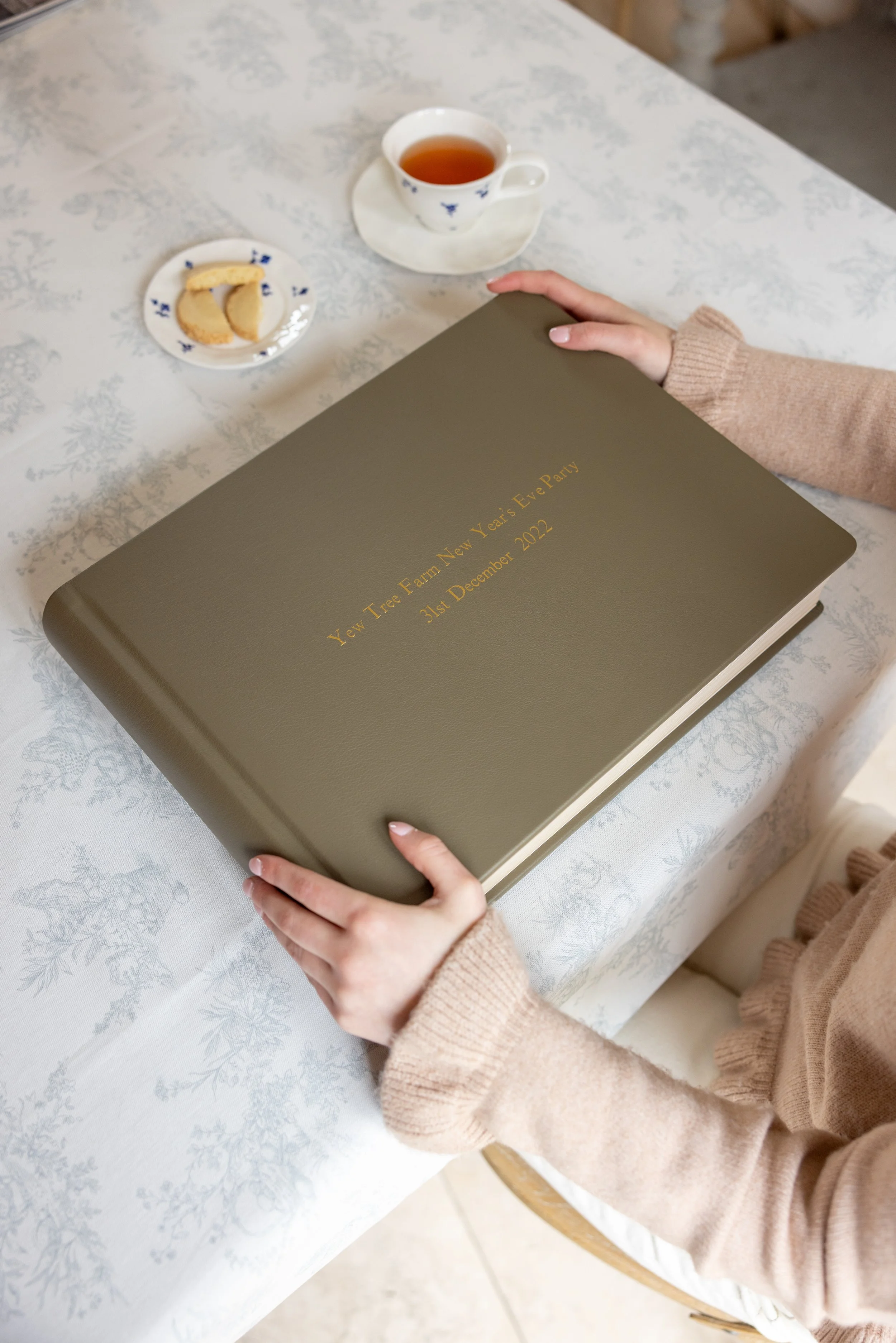 A person in a beige sweater holding a green guestbook titled 'Yew Tree Farm New Year’s Eve Party 31st December 2022' at a table with a tea cup, a small plate with cookies, and a white cloth with a silver woodland pattern.