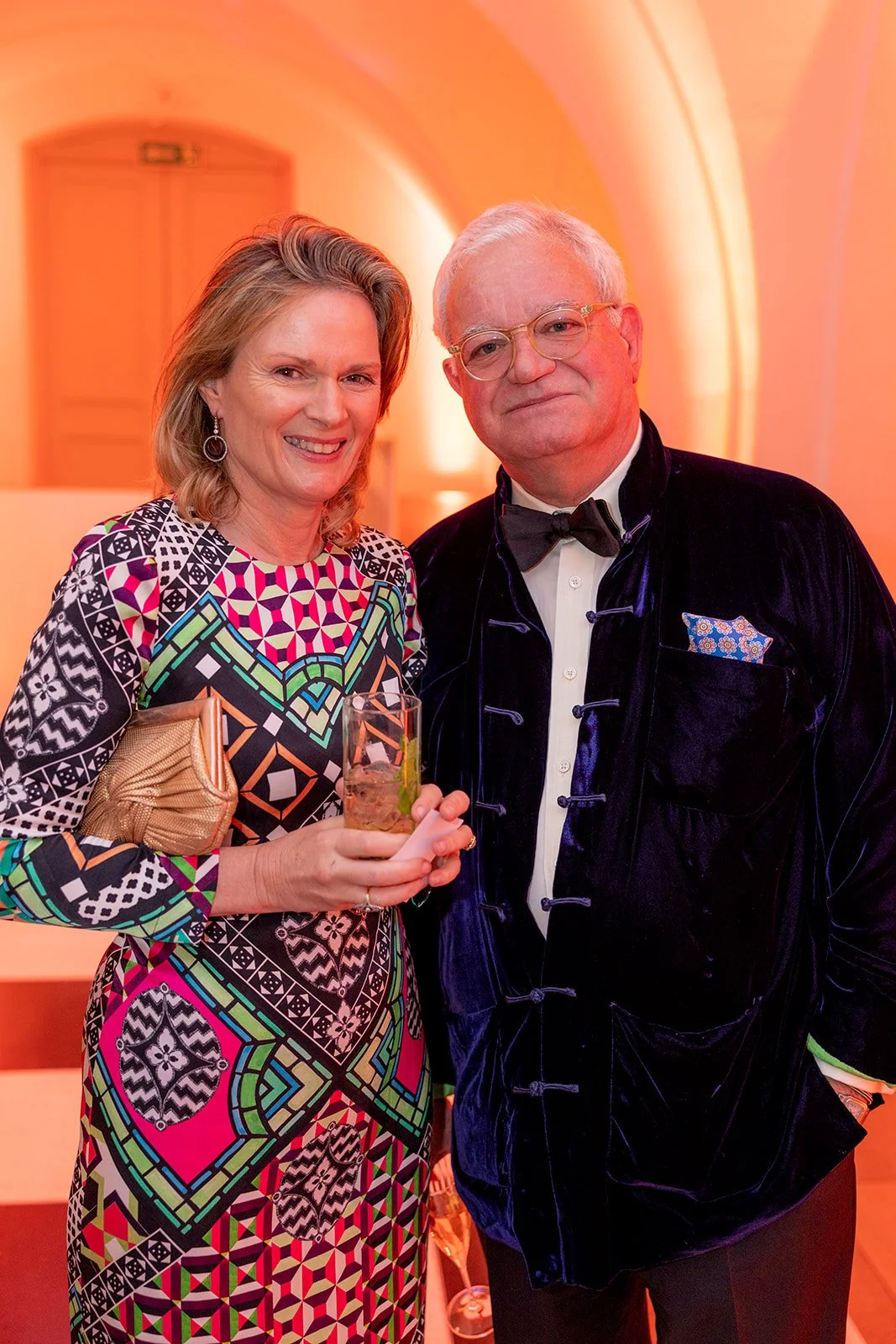A woman and a man dressed in formal attire at an indoor event, posing for a photo. The woman is holding a drink and wearing a colorful patterned dress with a gold clutch, smiling at the camera. The man is wearing a black velvet jacket, bow tie, and g