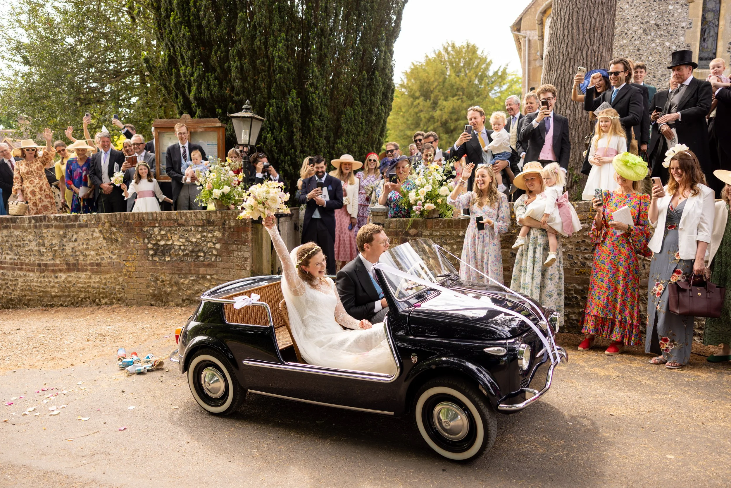A wedding celebration outdoors with guests gathered and taking photos, with a bride and groom in a vintage convertible car, and a group of people cheering, waving, and smiling.
