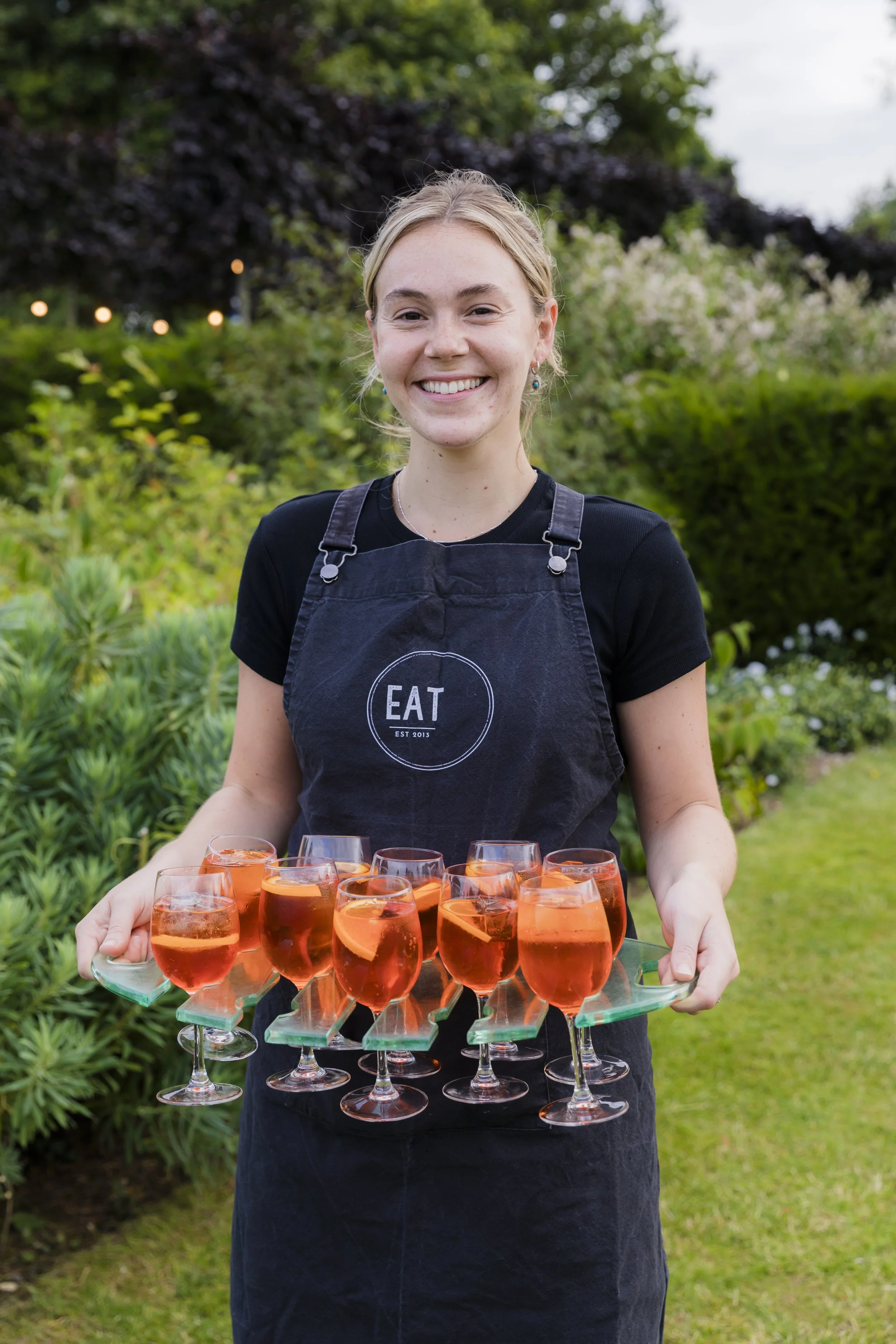 A smiling woman wearing a black apron with 'EAT' written on it, holding a tray of ten glasses filled with pink-colored drinks with lemon slices, outdoors in a garden.