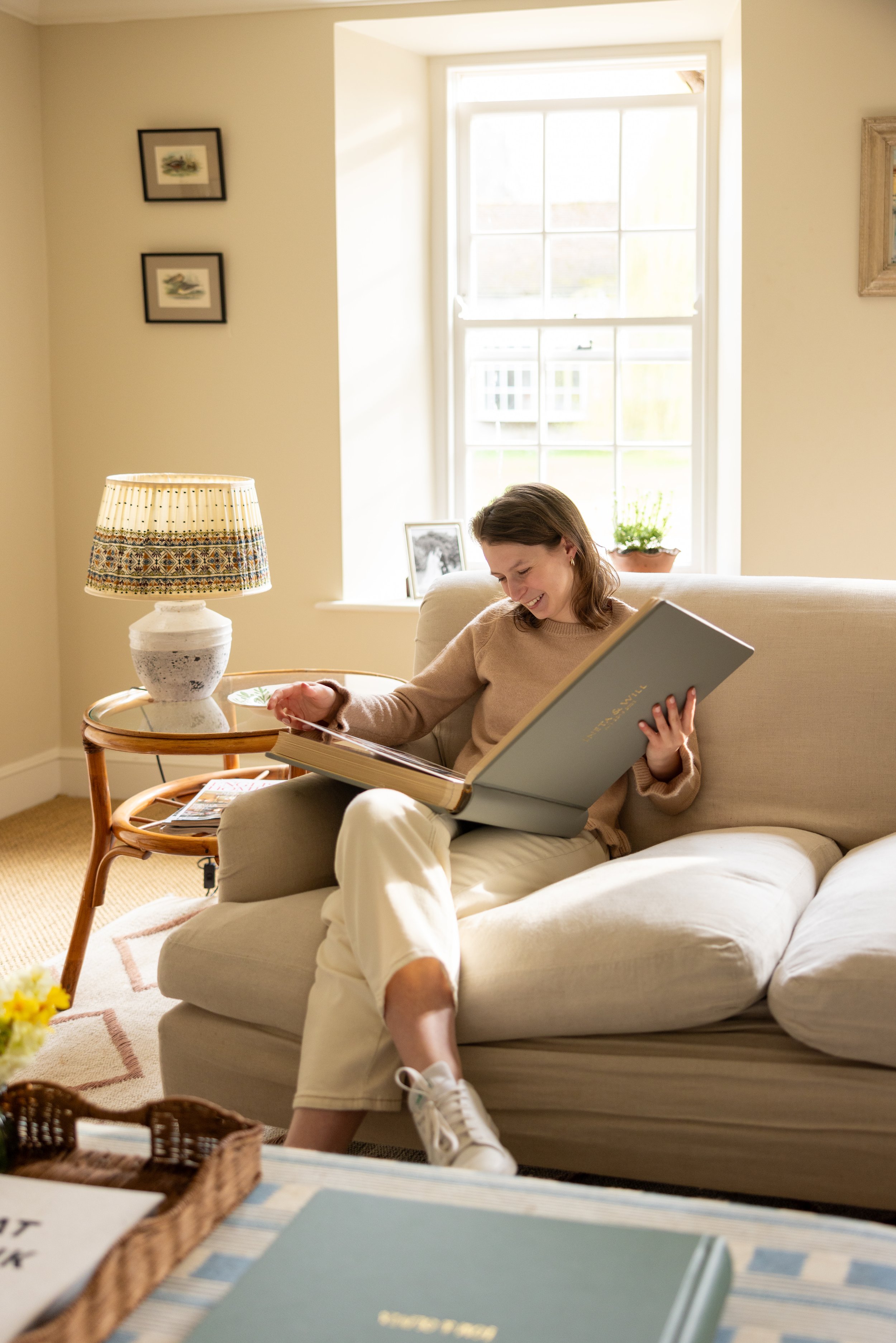 A woman sitting on a beige sofa, smiling, as she looks at a book. There is a window behind her with bright light coming in, a small framed photograph and a potted plant on the windowsill. A patterned table lamp is beside her on a side table.