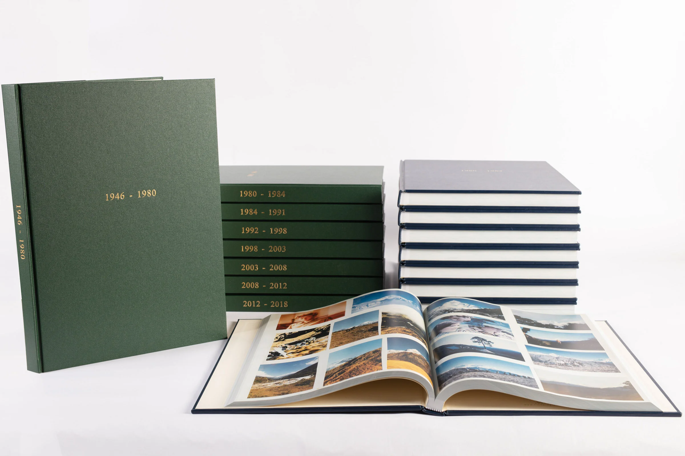 Stack of green photo albums labeled with years and a blue photo album open showing landscape photos, all on a white background.