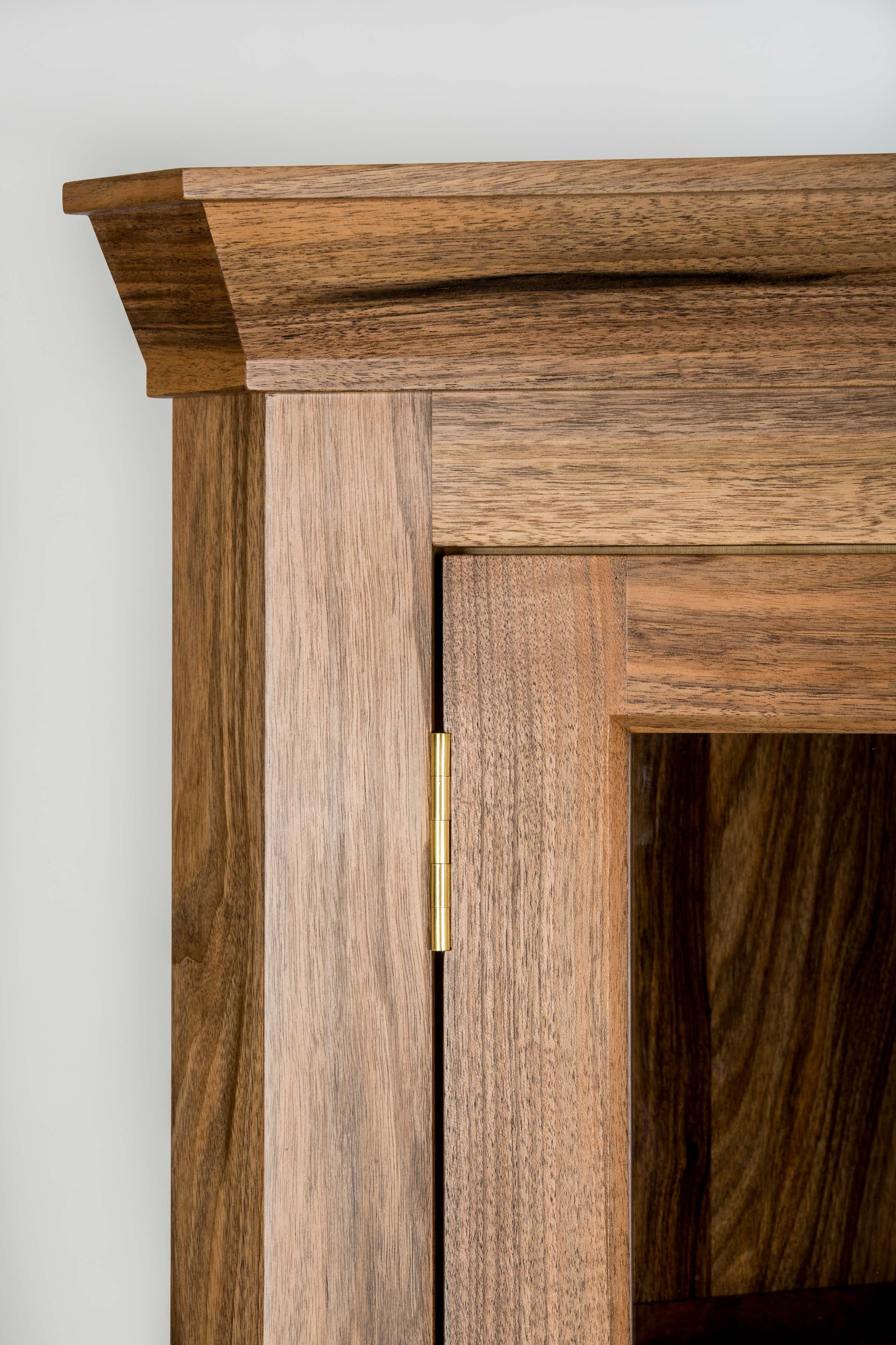 Close-up of a wooden door frame with a brass hinge, showing the top corner against a white wall.