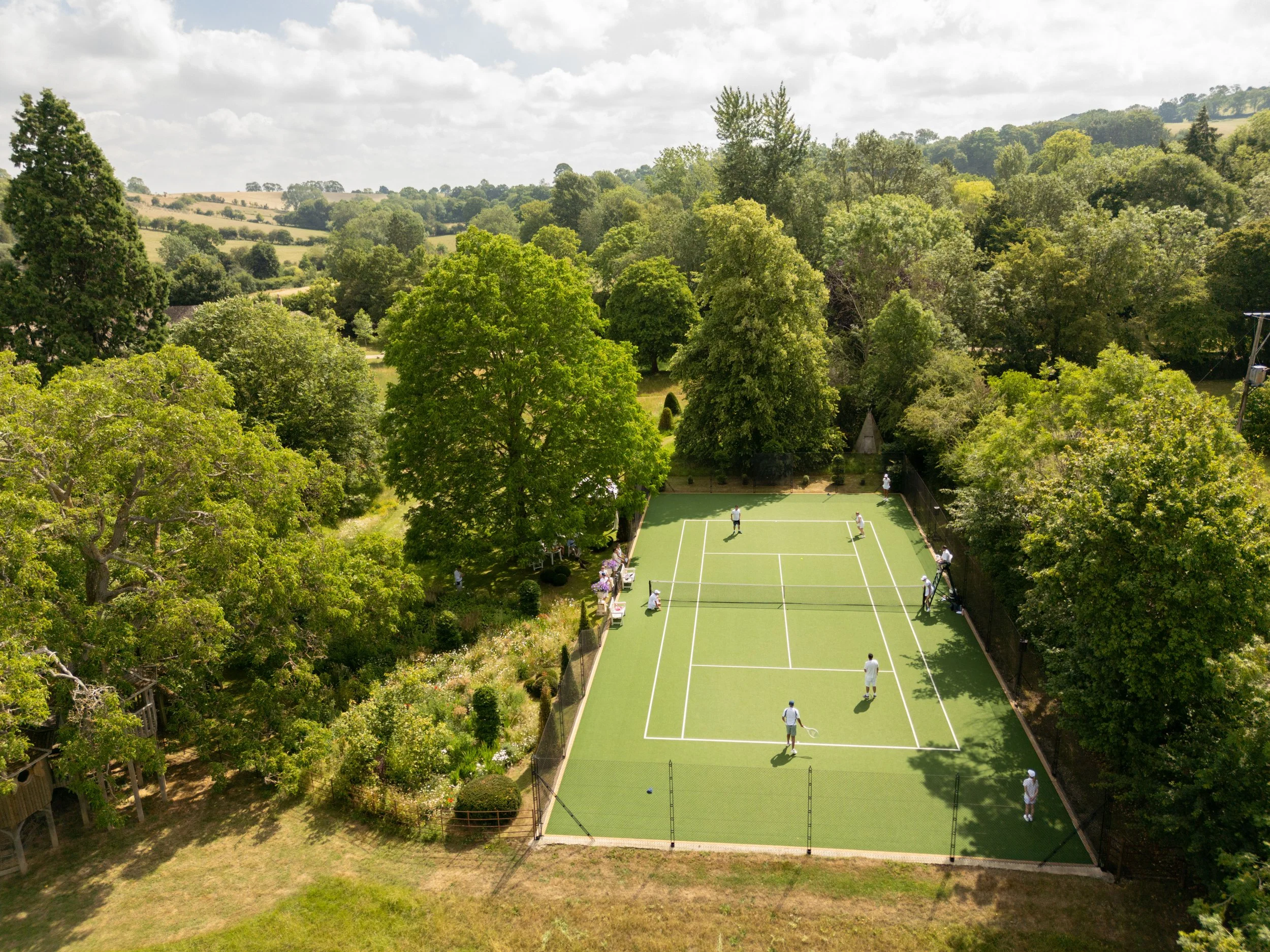 An aerial view of a tennis court surrounded by trees, with several people playing and standing on the court in sunlight.