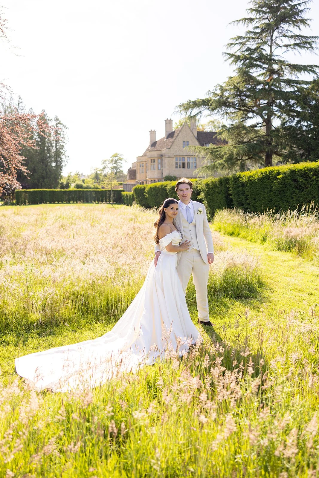 A bride and groom in elegant wedding attire standing in a grassy field with wildflowers, a large house and trees in the background on a sunny day.