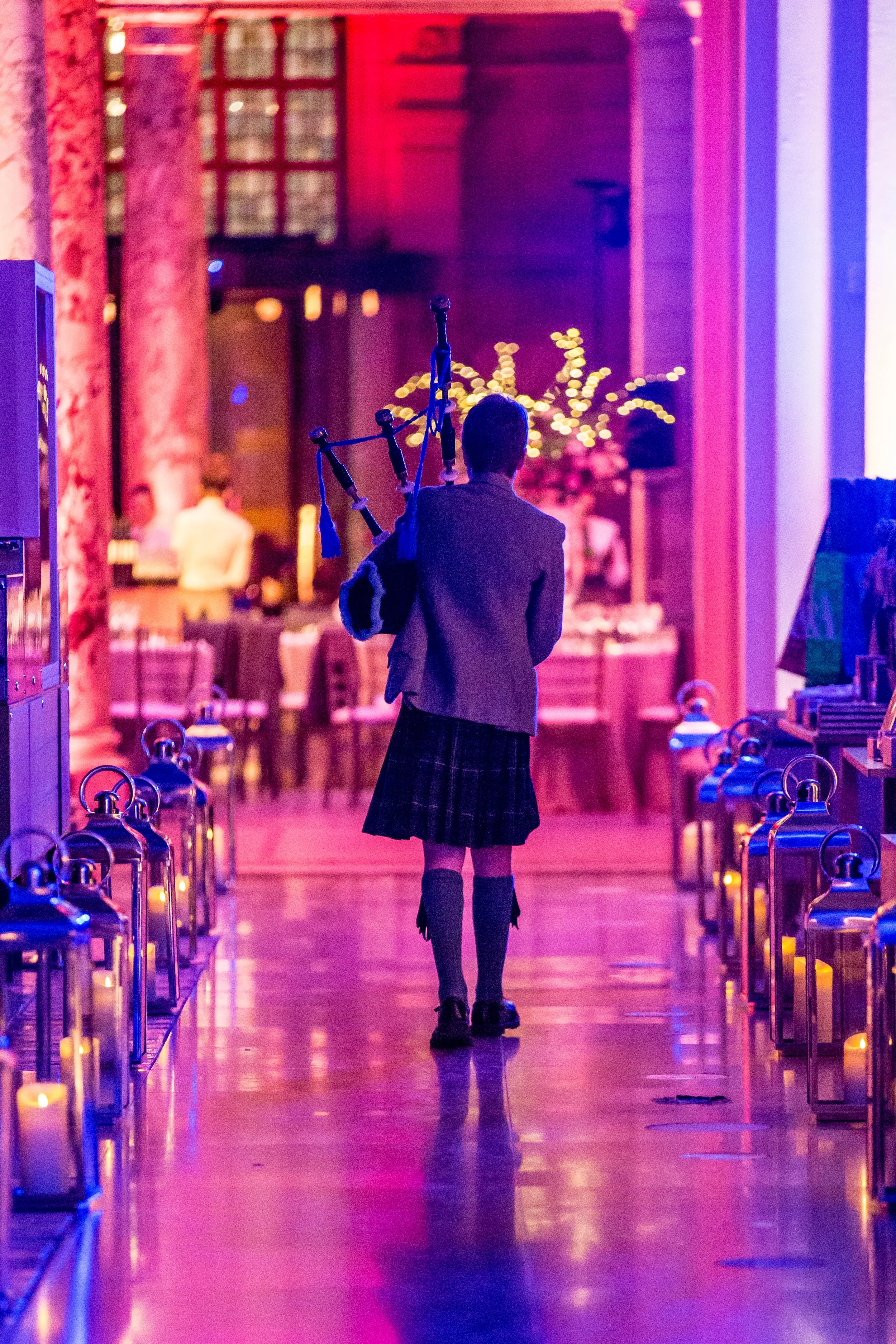 Person walking with bagpipes in an indoor event space decorated with warm lighting, tables, chairs, and floral arrangements.