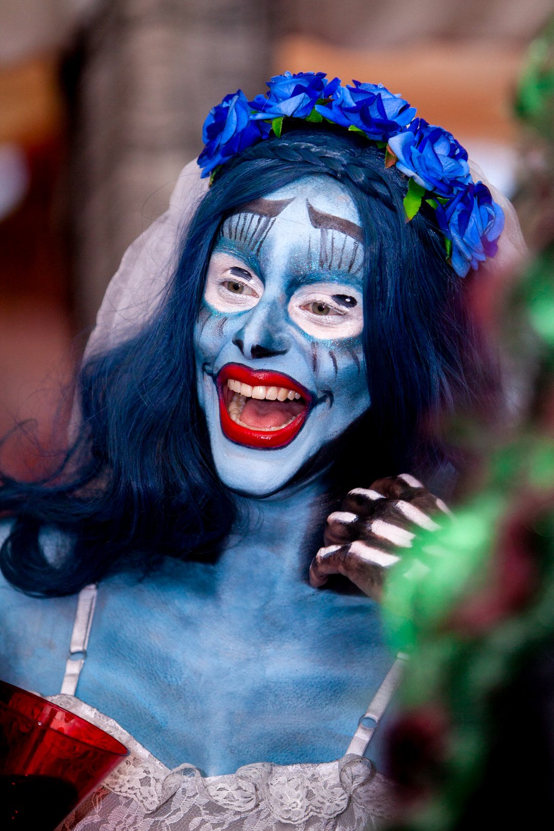 Person with blue face paint, red lipstick, and long dark hair wearing a flower crown, mid-laugh, in costume for a celebration or event.