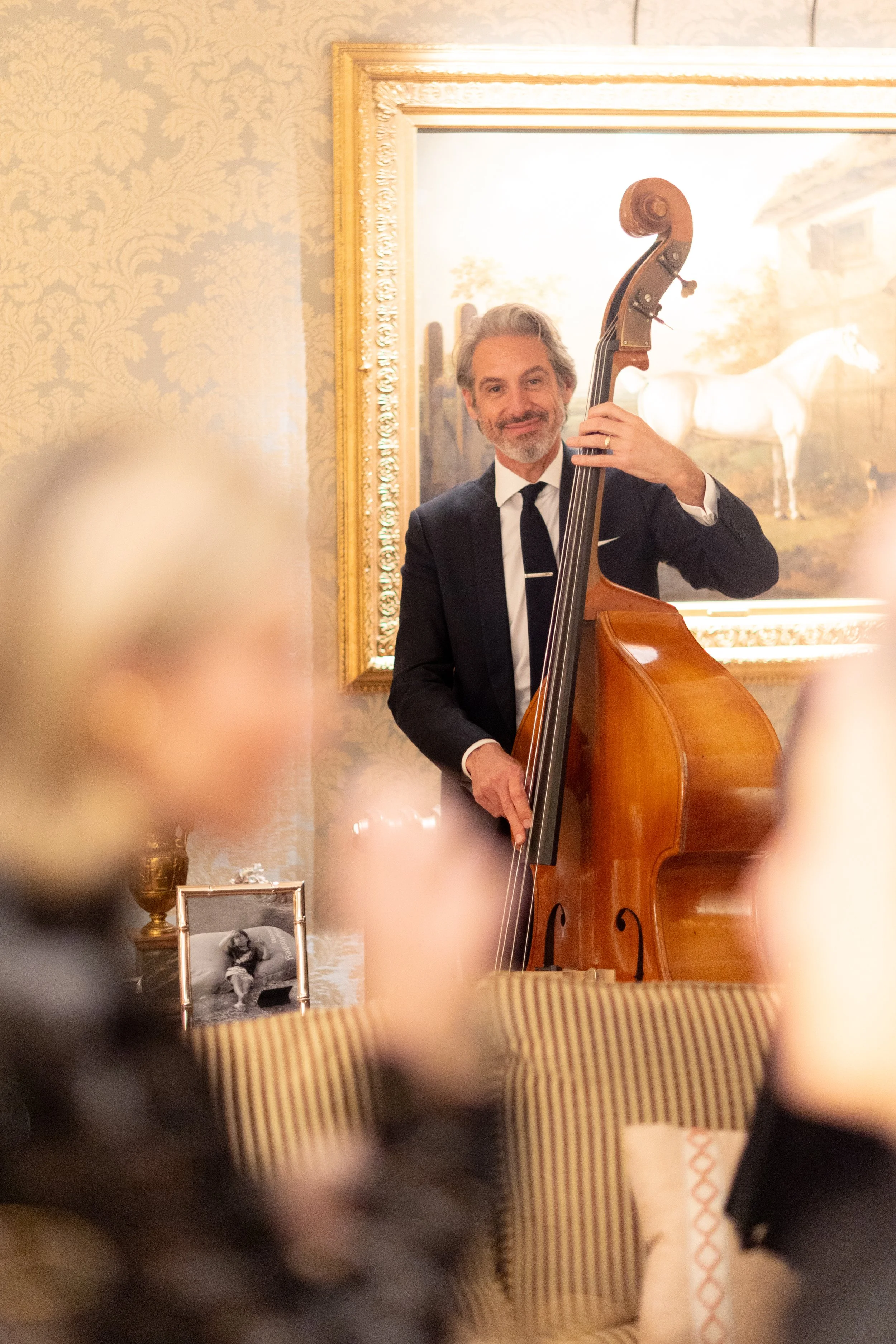 A man in a black suit playing a double bass at a social gathering in an ornately decorated room with a framed painting of a horse on the wall behind him.