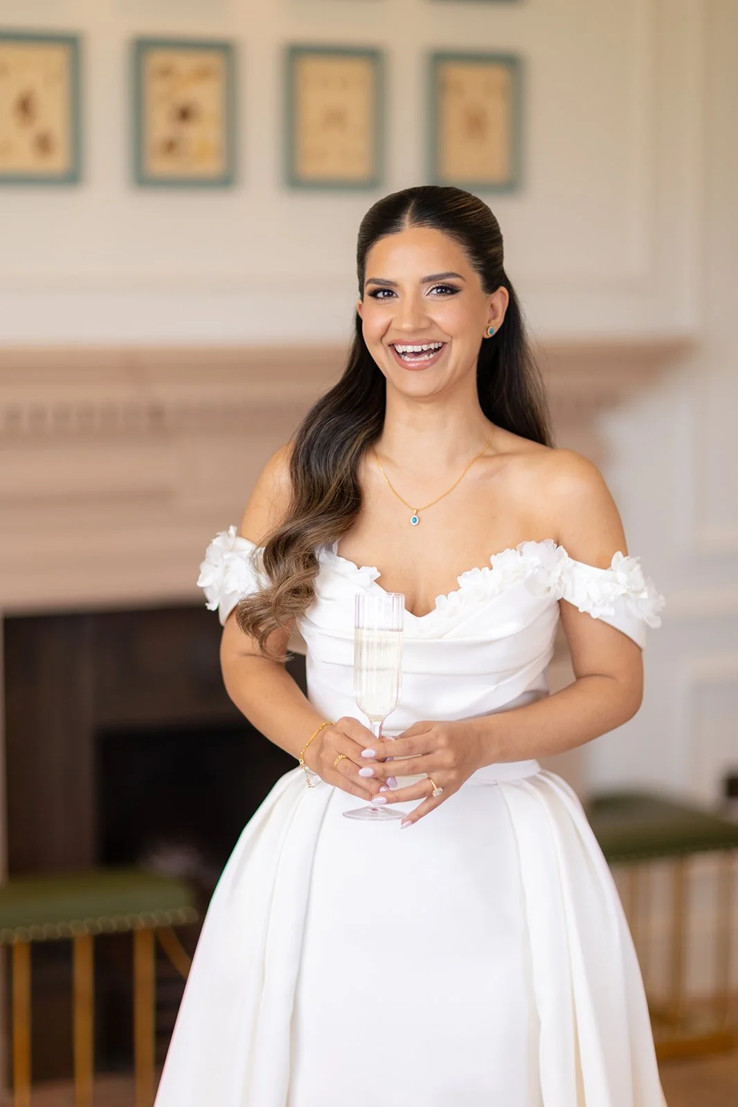A woman in a white off-the-shoulder wedding dress smiling and holding a champagne flute, standing indoors with framed artwork on the wall behind her.