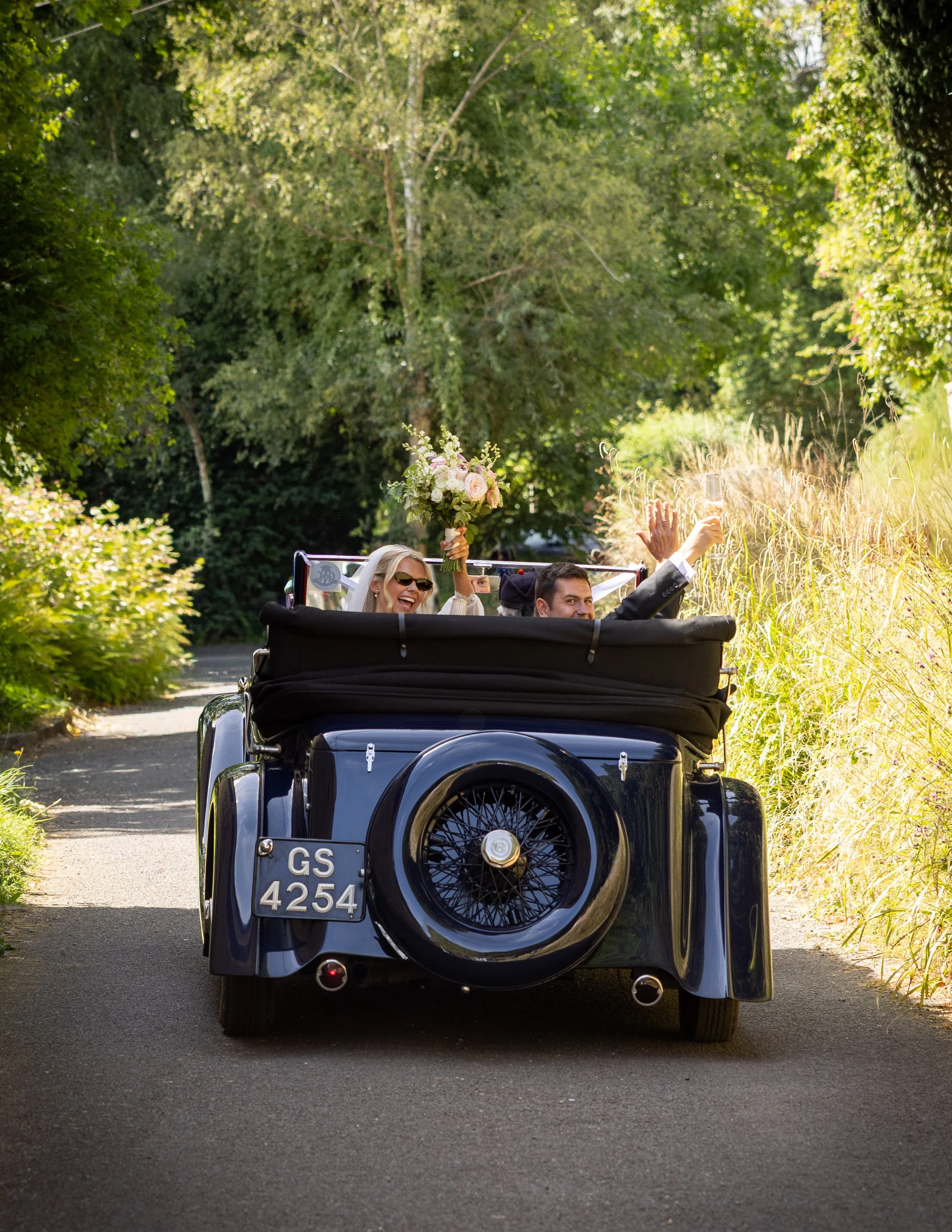 The back view of a vintage black car with a spare tire mounted on the rear, surrounded by lush green trees and tall grass along the roadside, with a woman holding a bouquet of flowers and waving, and a man sitting beside her, both smiling.