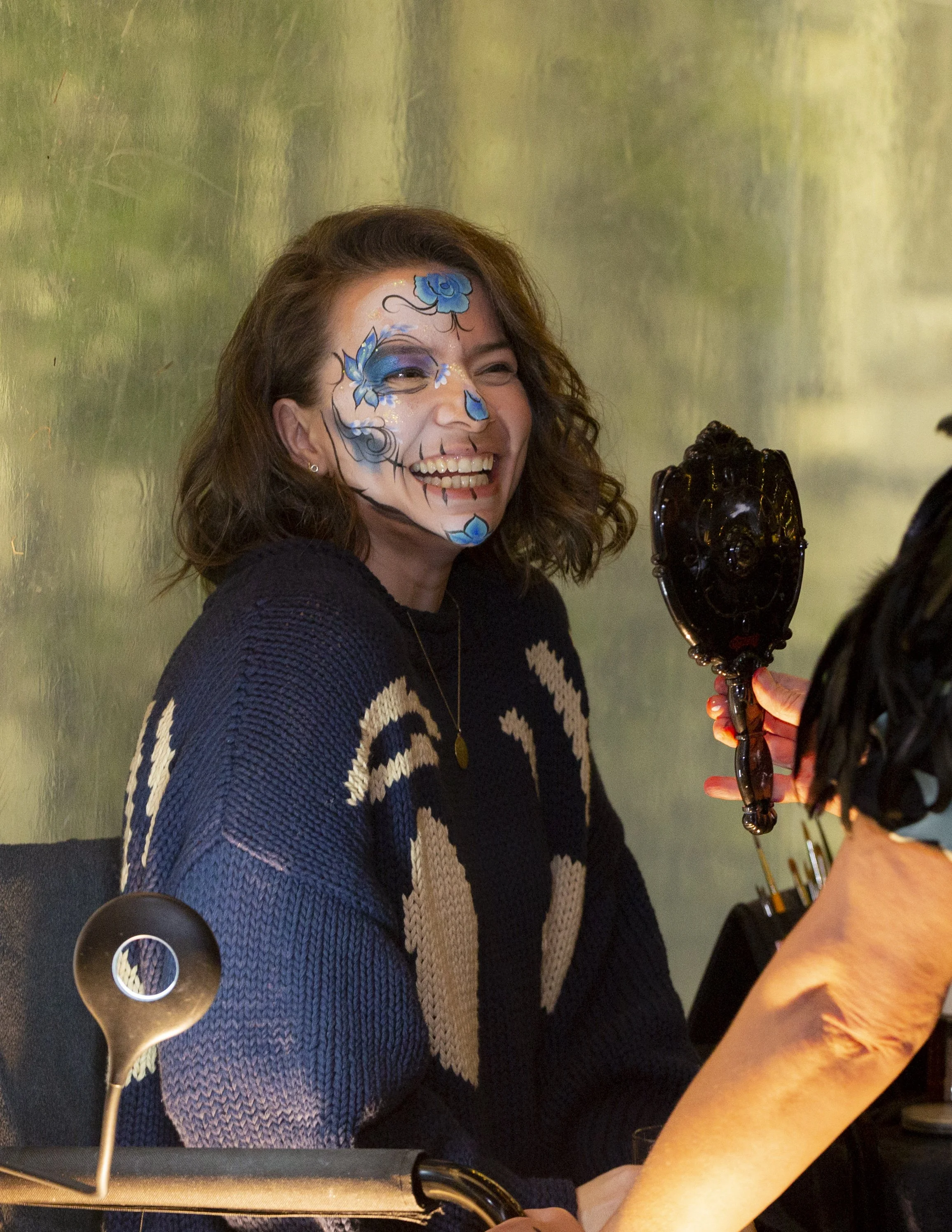 A woman with curly brown hair smiling as she gets her face painted with blue floral designs.