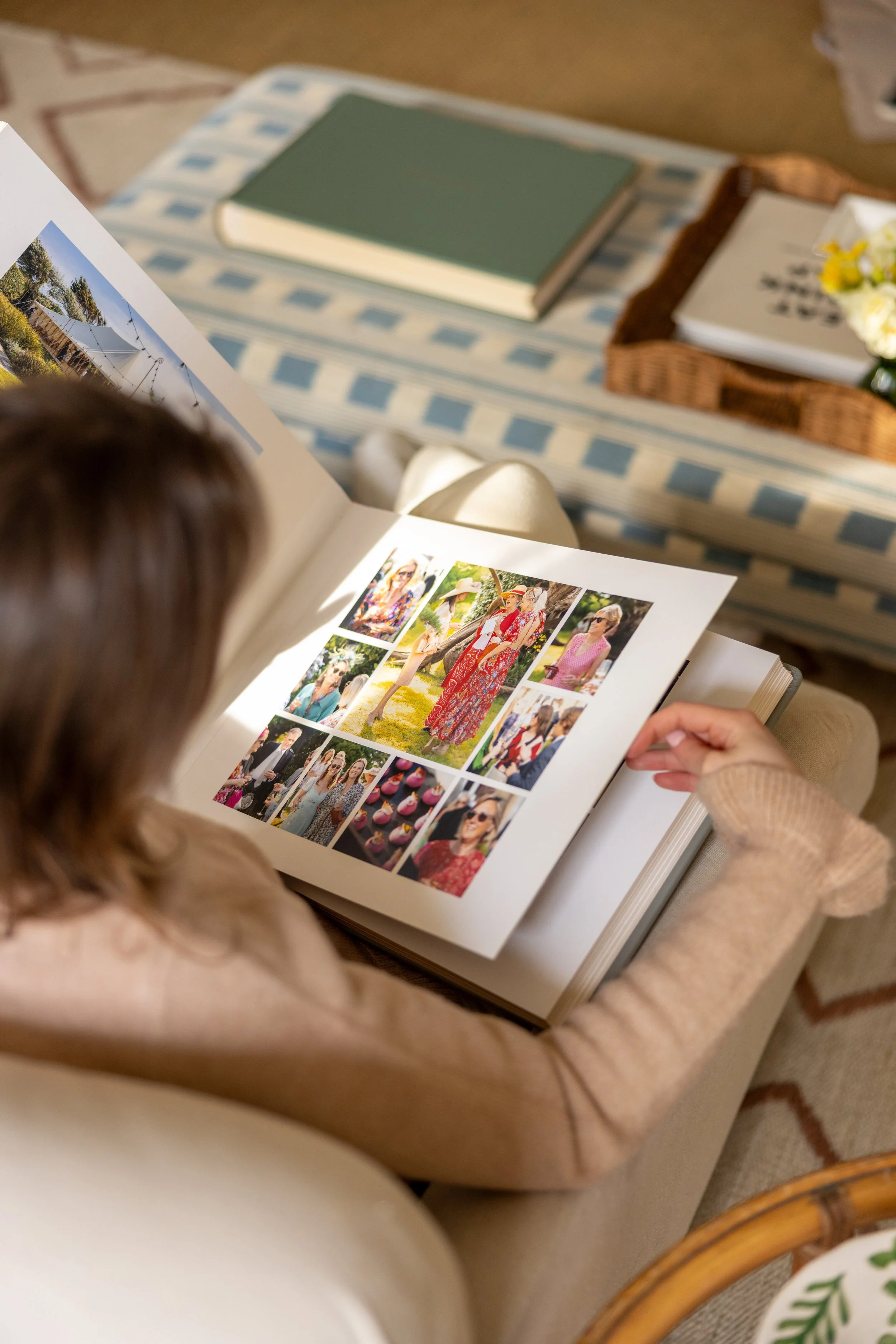 A person looking through a photo album with pictures of people at outdoor events, including individuals in hats and colorful clothing, as well as photos of cupcakes. The background shows a striped blue and white ottoman with a book and a wicker tray 