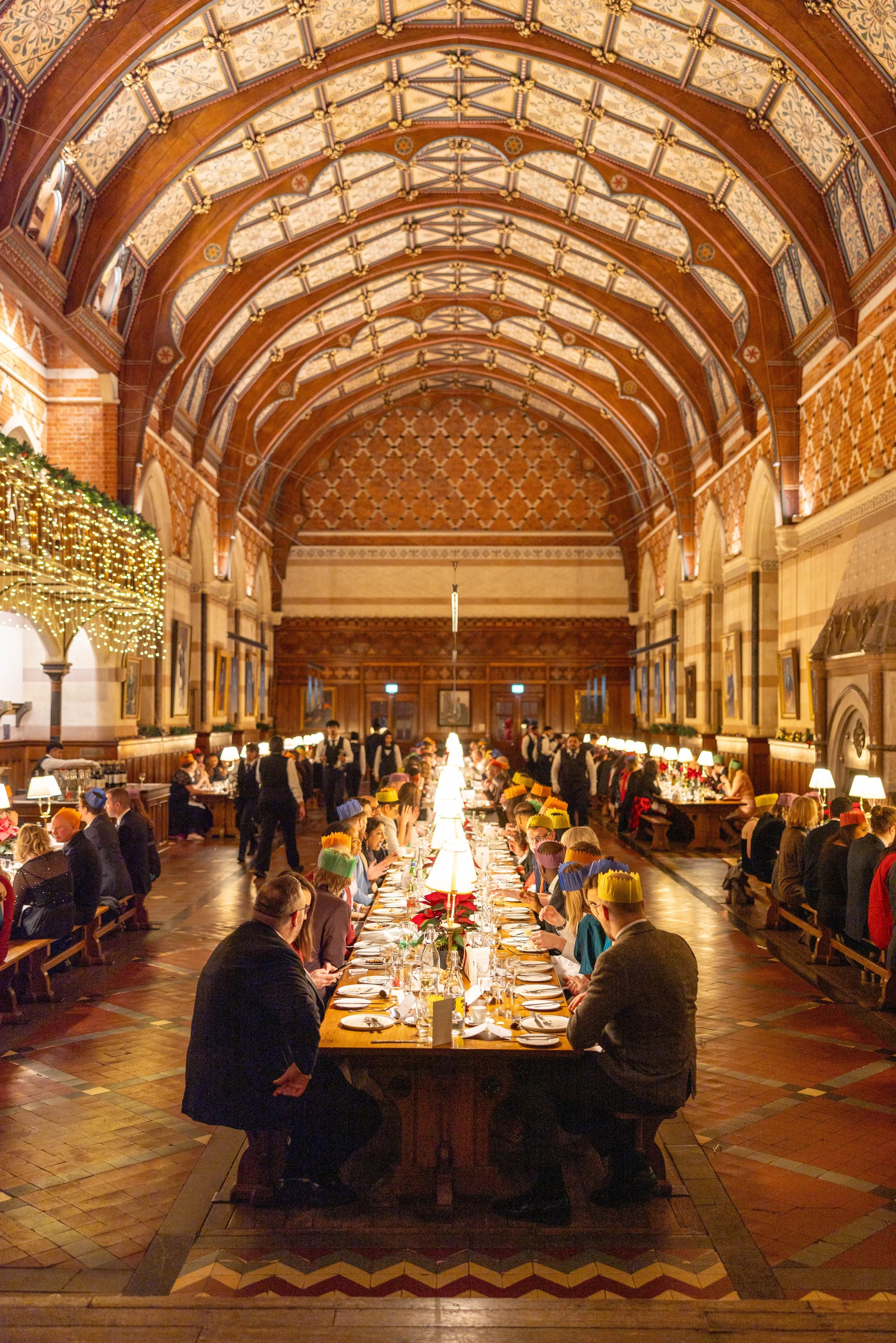 A large, elegant dining hall decorated for a festive event with Christmas lights and ornaments, featuring a long banquet table set with plates, glasses, and candles, and people wearing colorful paper crowns seated around it.