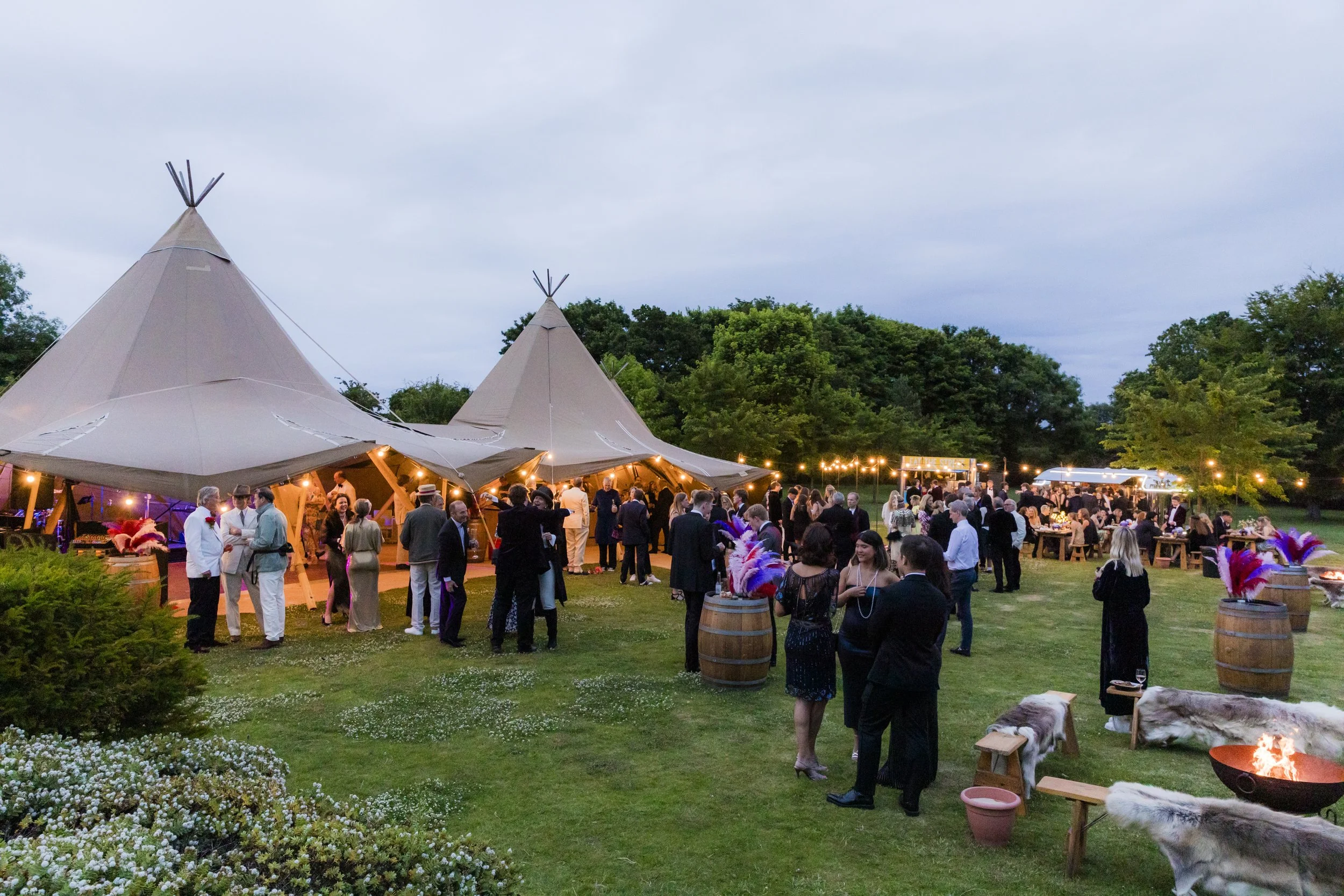 Outdoor gathering with people socializing and dining at a festive event in a large open area with teepees, string lights, and decorated barrels, during early evening.