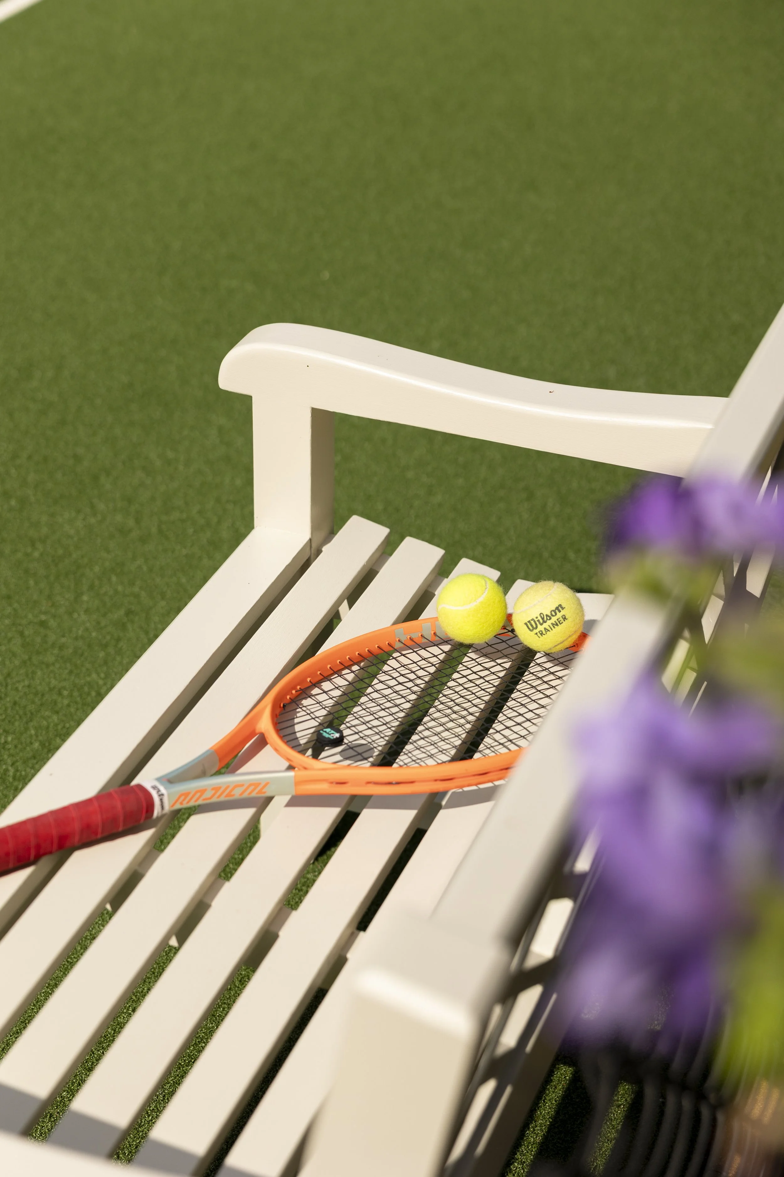 Tennis equipment including a tennis racket, two tennis balls, and a small charm on a white bench with a green artificial grass surface in the background.