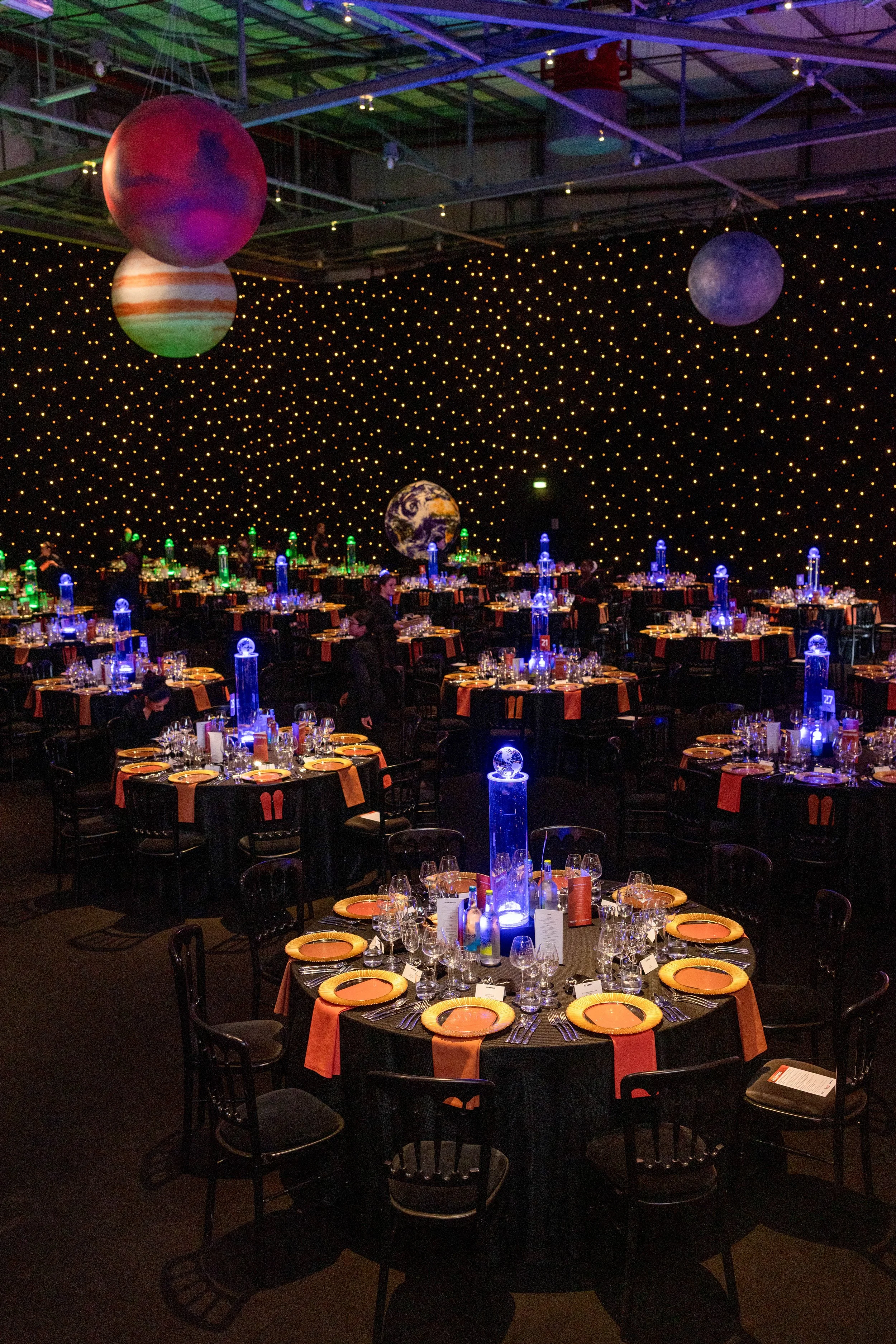 Event space with space-themed decorations, including planets and stars hanging from the ceiling, and tables set with elegant place settings and illuminated centerpieces.