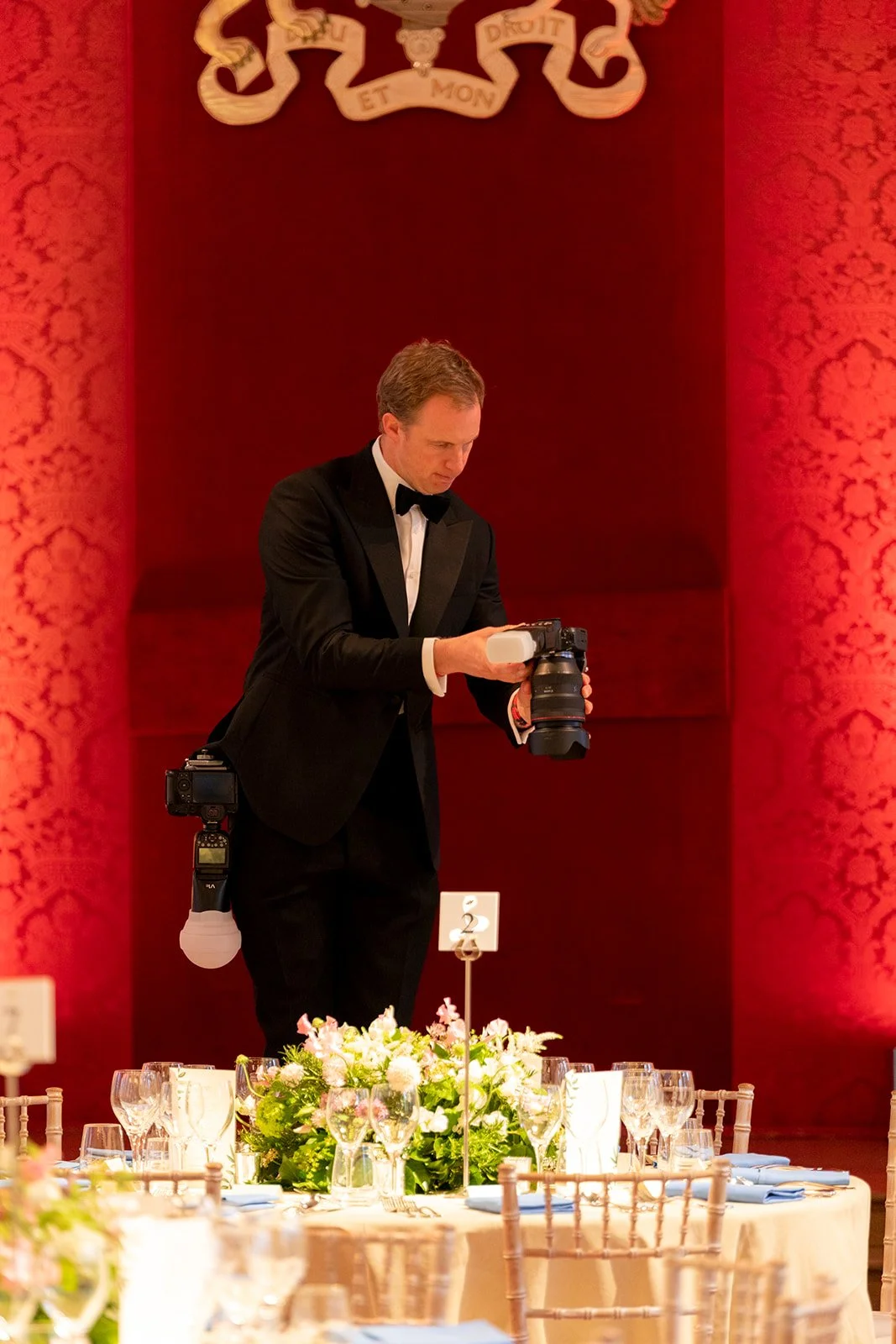 A man in a tuxedo setting up a camera on a tripod at a formal event with a decorated table and floral centerpiece in the foreground, and ornate red wallpapered walls with a crest or sign overhead.