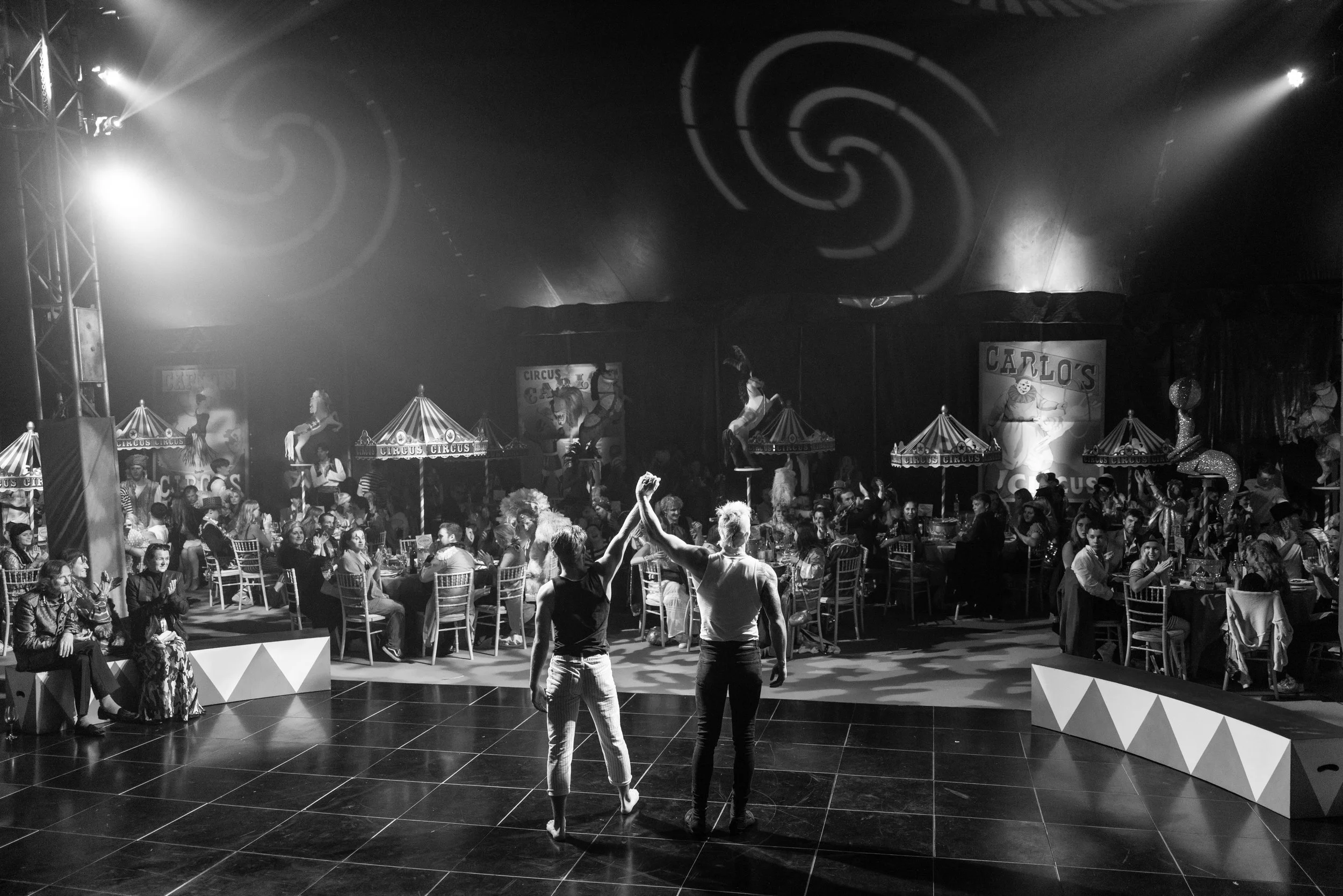 Circus performance with two acrobats dancing on stage, surrounded by an audience seated at tables in a large tent with decorative lighting and circus-themed decor.