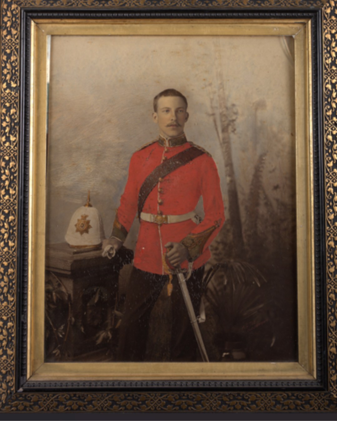 Portrait of a young man dressed in military uniform with a red coat, gold accents, and a black sash, standing next to a table with a white helmet featuring a gold emblem, holding a sword, with a blurred outdoor background.