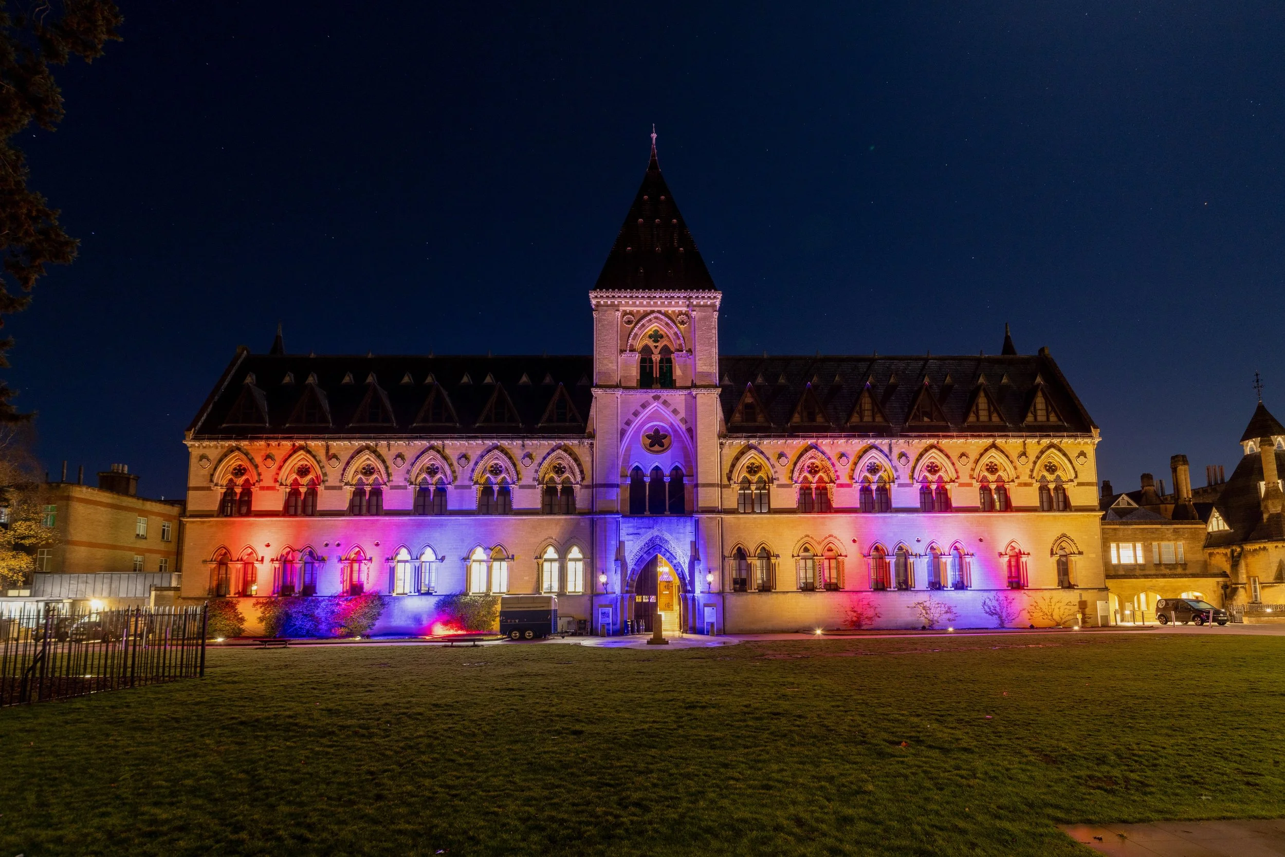 Night view of a historic stone building illuminated with colorful purple, pink, and red lights, featuring Gothic windows and a central tower with a pointed roof, set against a starry sky.