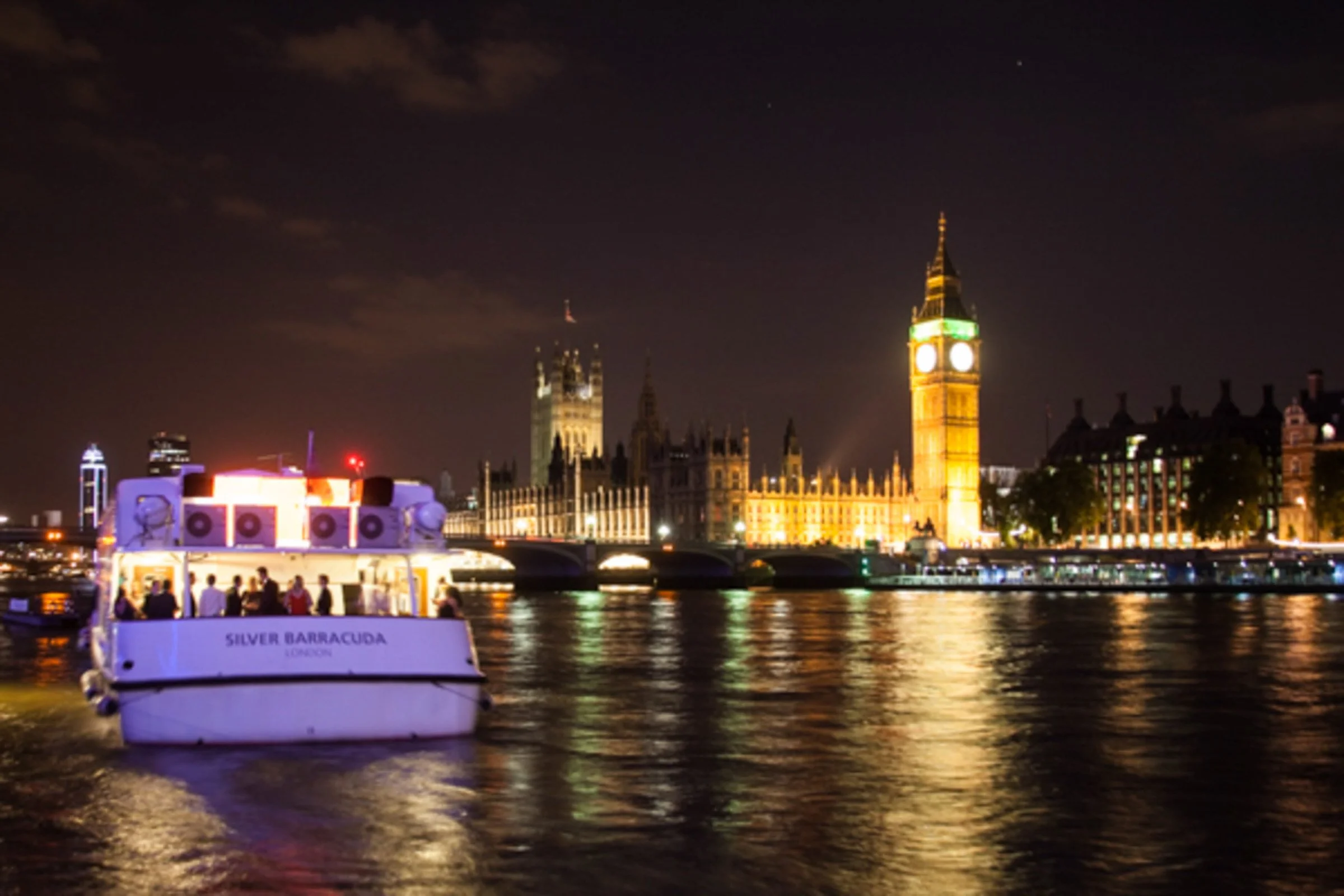 Nighttime view of London skyline with illuminated Big Ben and government buildings, with a boat on the River Thames.