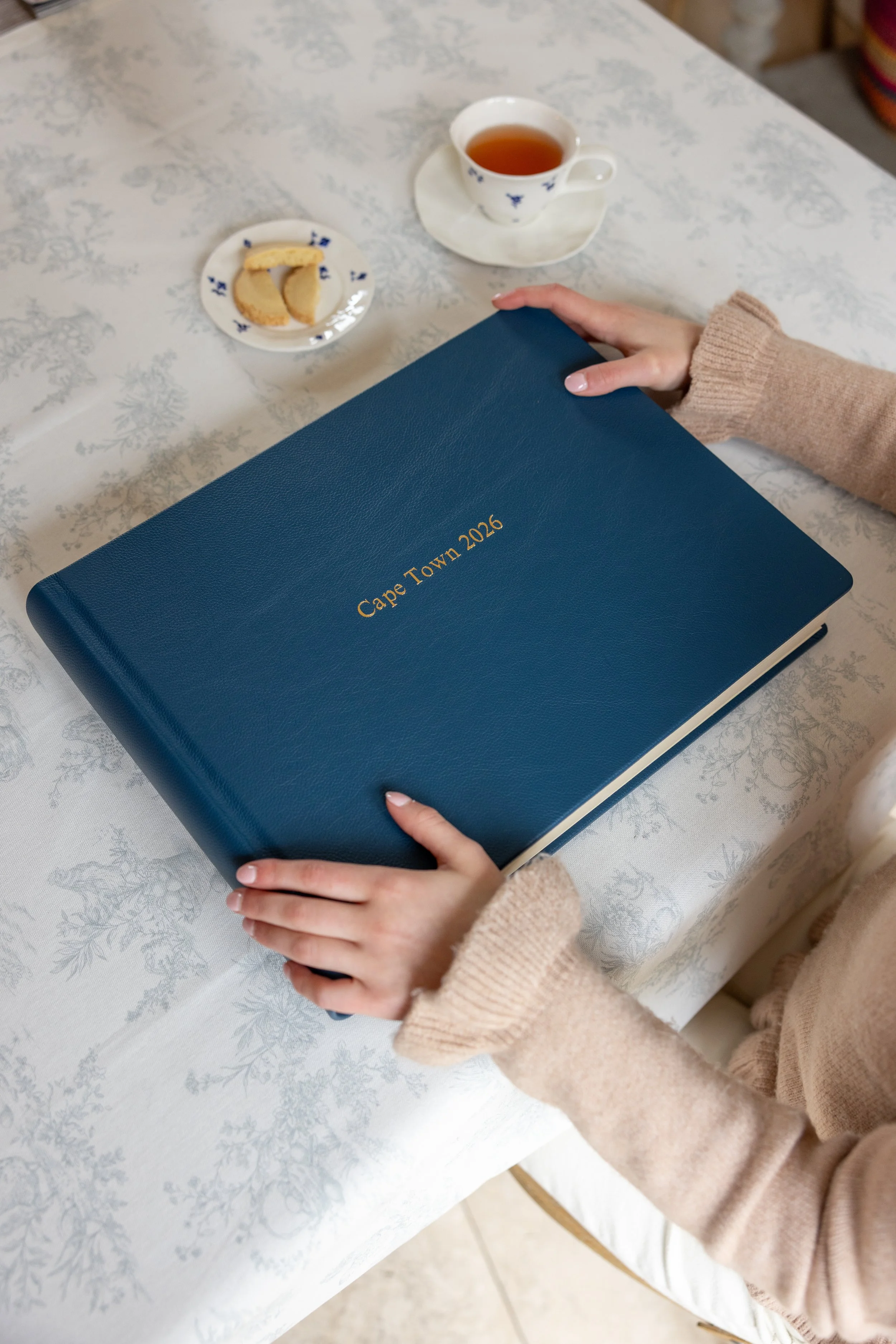 A person holding a blue planner titled 'Cape Town 2026' sits at a table with a cup of tea and a small plate of cookies.