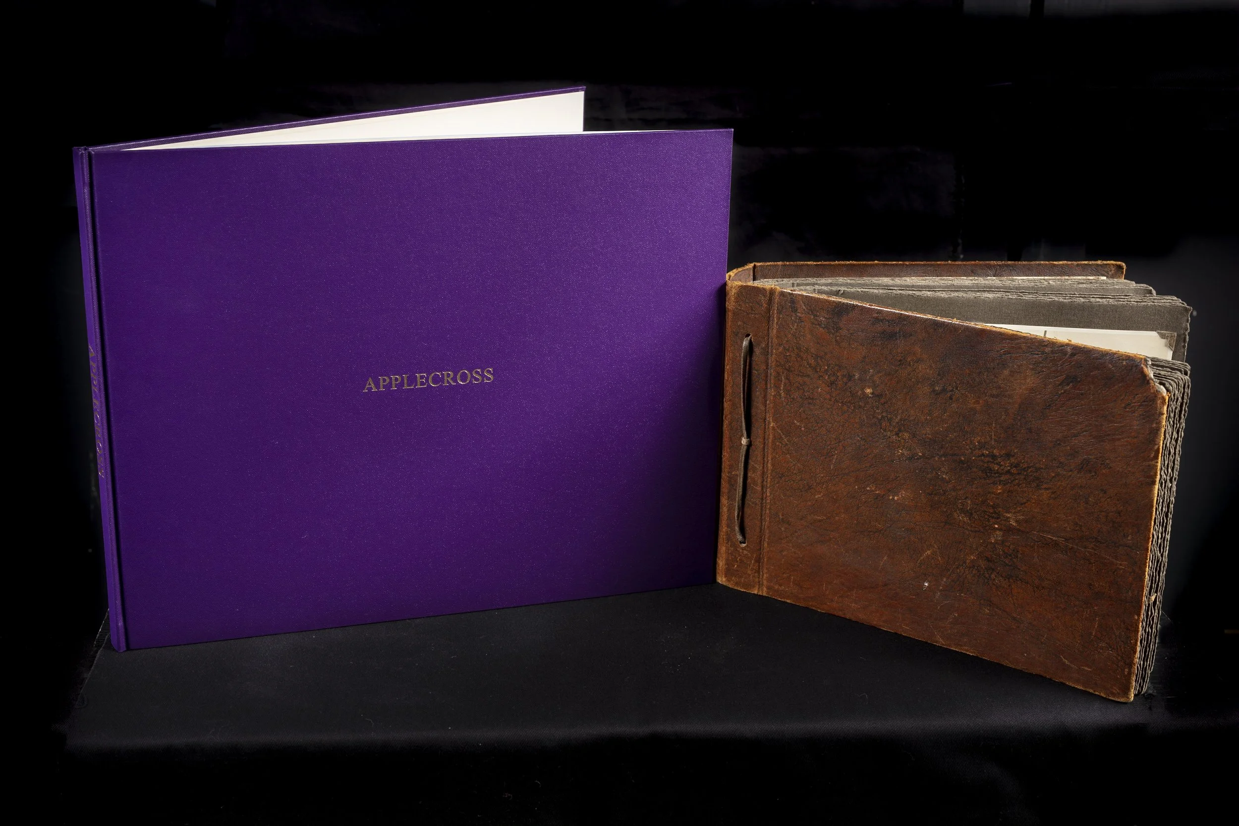 Two books side by side on a black surface, one purple with gold lettering saying 'APPLECROSS' on the cover, and the other brown with a textured leather-like cover