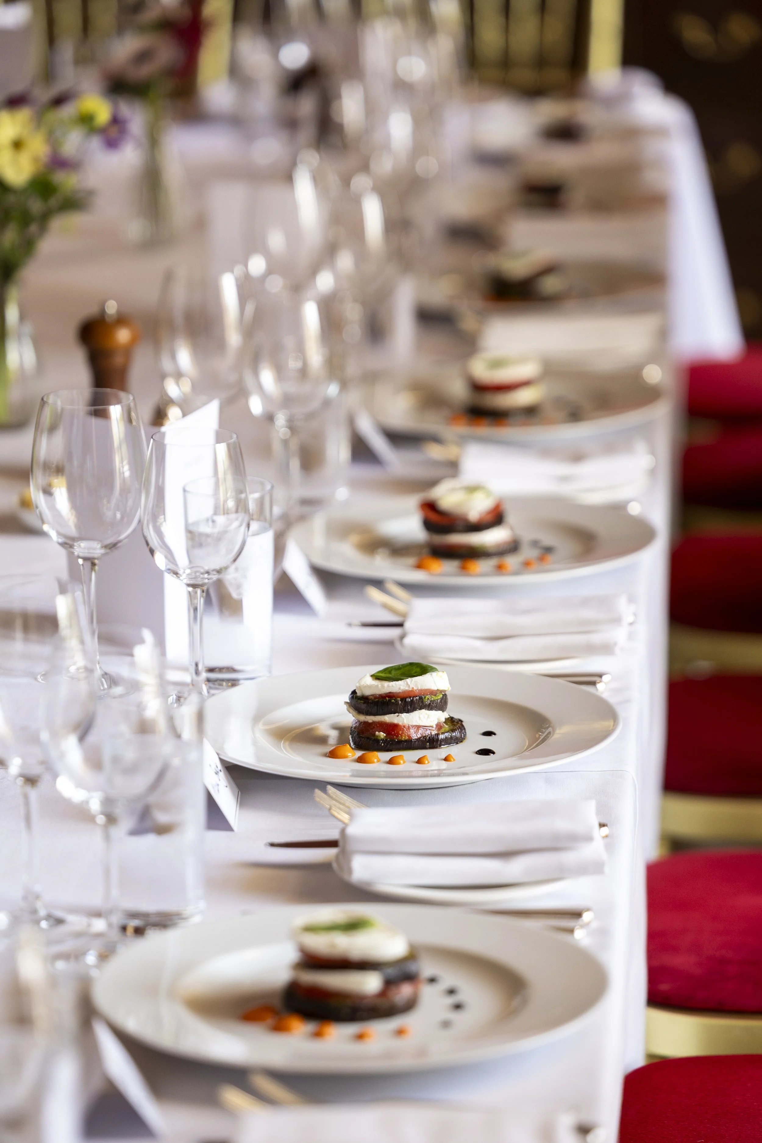 A formally set dining table with plates of appetizer or small dish, wine glasses, water glasses, cutlery, and white napkins, in a restaurant or banquet setting.